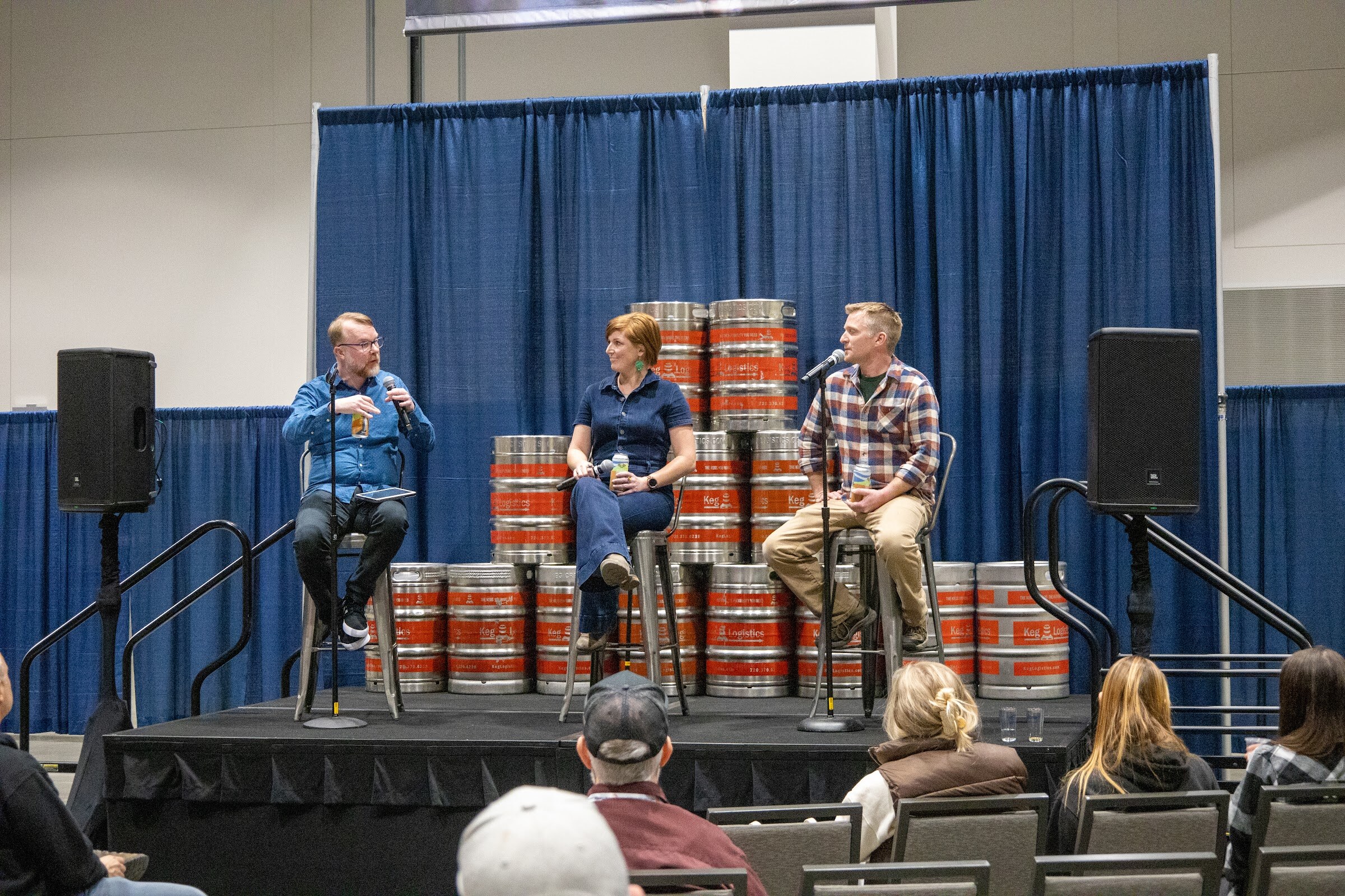Three panelists seated on stools in front of stacked kegs on a stage with a small audience watching