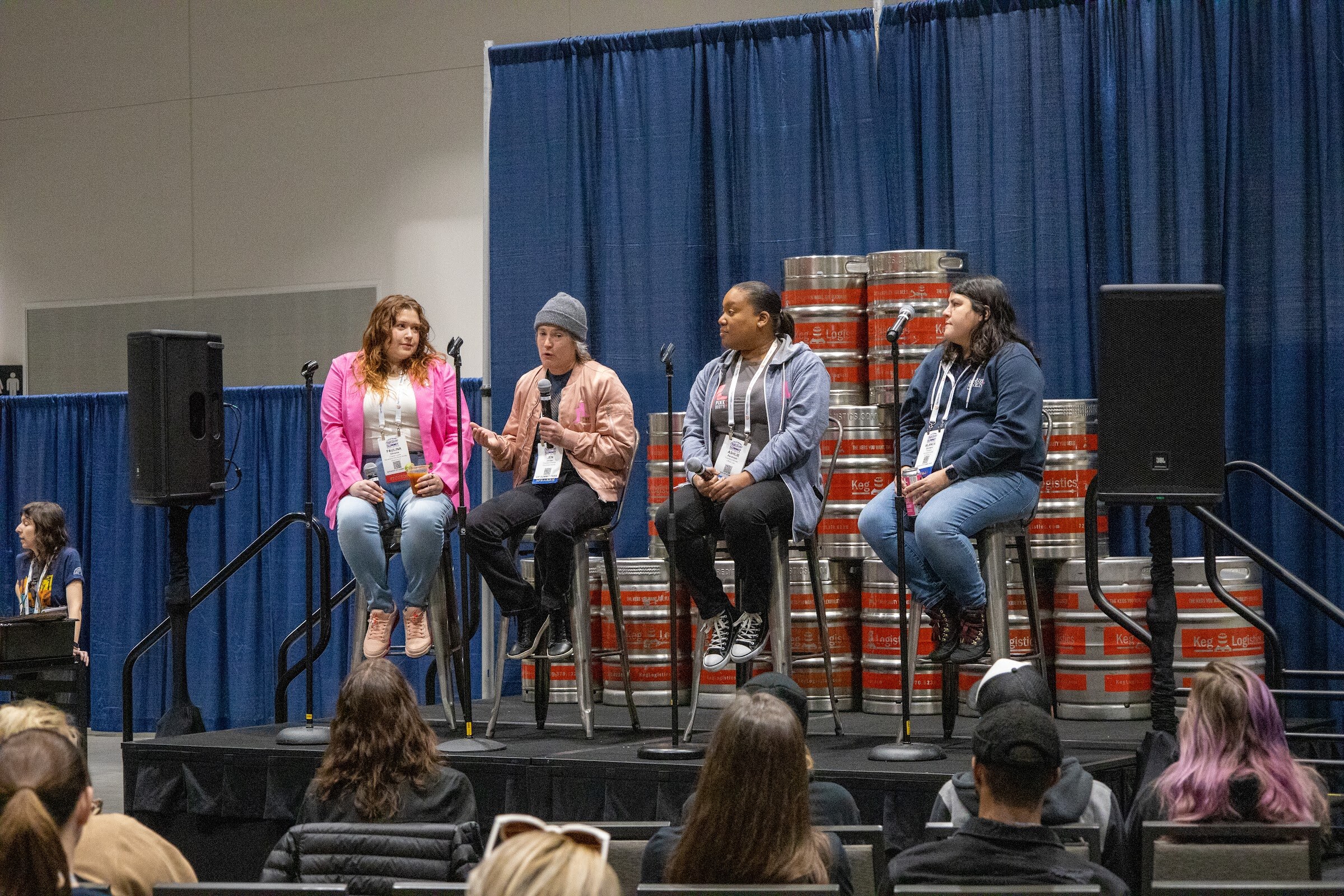 Four panelists seated on stools in front of stacked kegs under a blue curtain with attendees seated in front