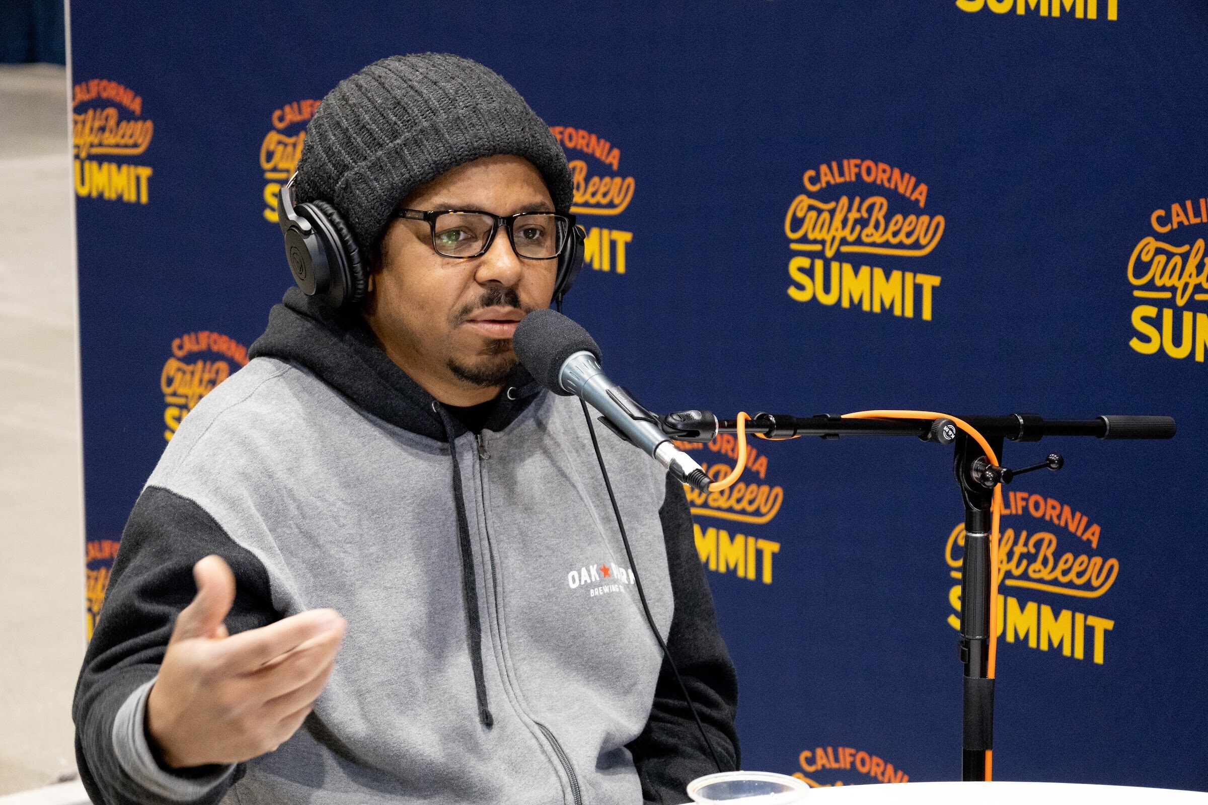 A man in a gray beanie, glasses and a hoodie gestures while speaking into a microphone in front of the California Craft Beer Summit backdrop