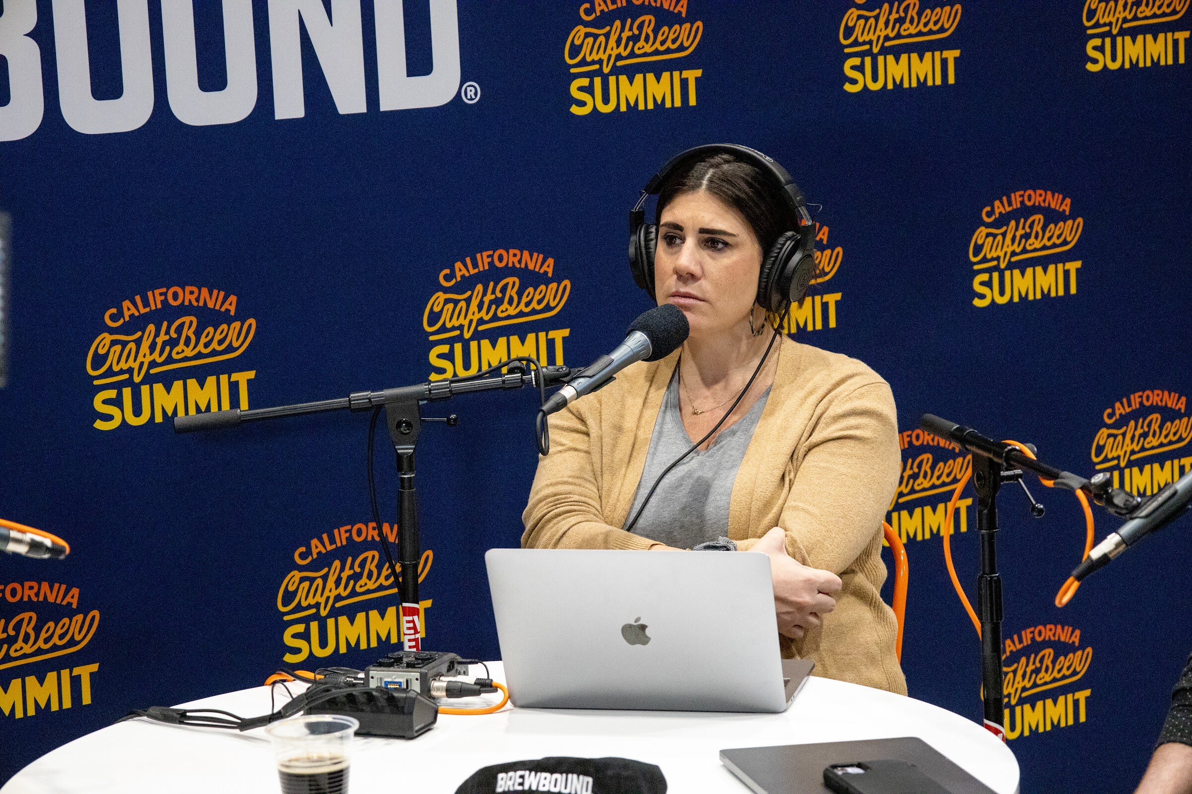 A woman in headphones and a tan cardigan sits at a table with a laptop and a Brewbound cap in front of the California Craft Beer Summit backdrop