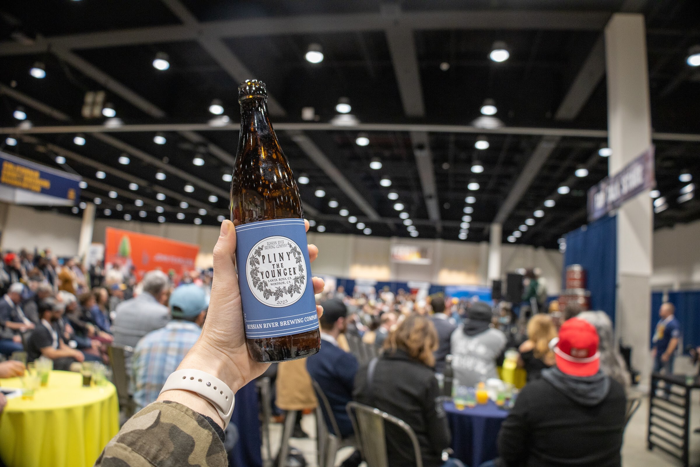 A hand holds up a bottle with a blue label in the foreground while a large crowd sits at round tables in the convention hall behind it