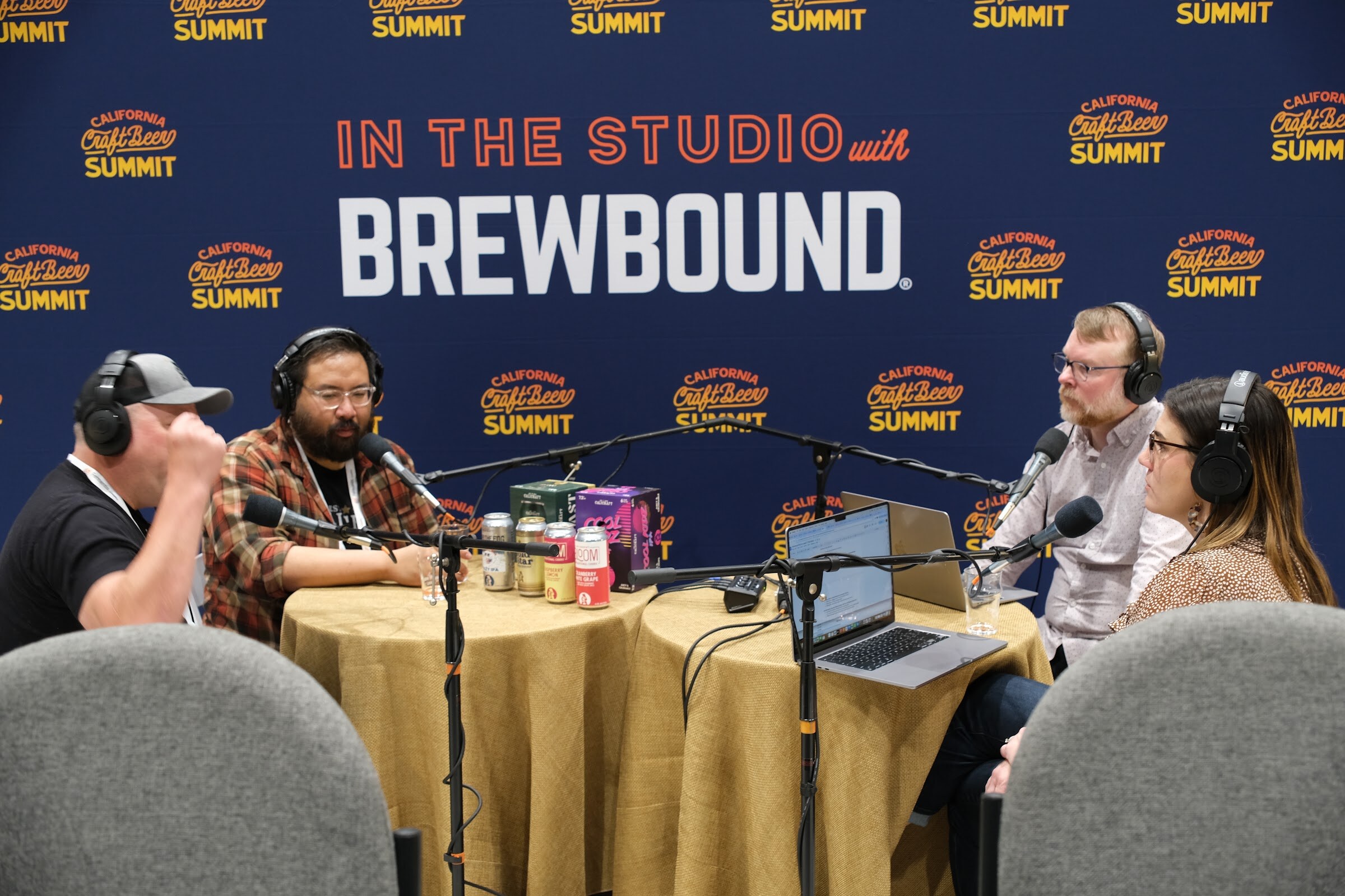 Two guests in headphones talk at a round table with beer cans in front of an In the Studio with Brewbound backdrop