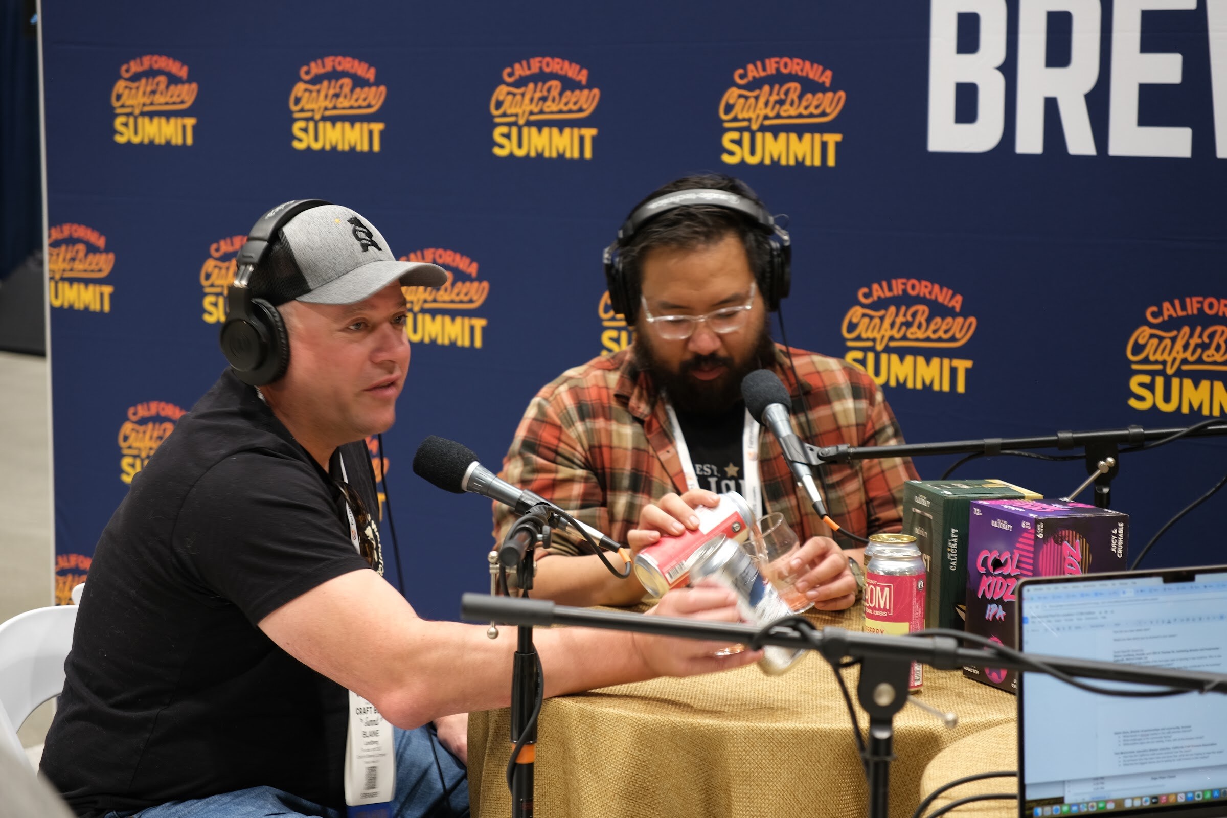 A man in a black T-shirt pours a canned beer into a glass at the podcast table while a second guest with a headset looks on