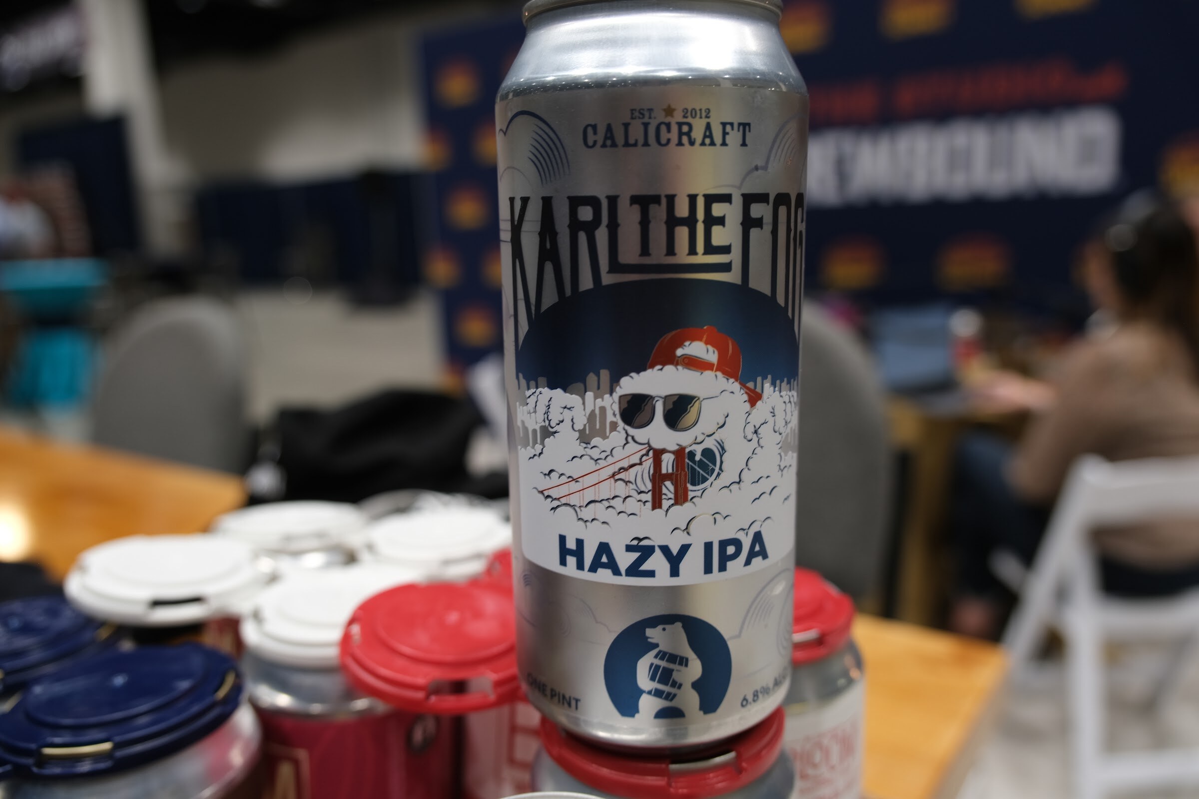 A silver Calicraft Karl the Fog Hazy IPA can in focus on the podcast table with blurred cans and cups behind it