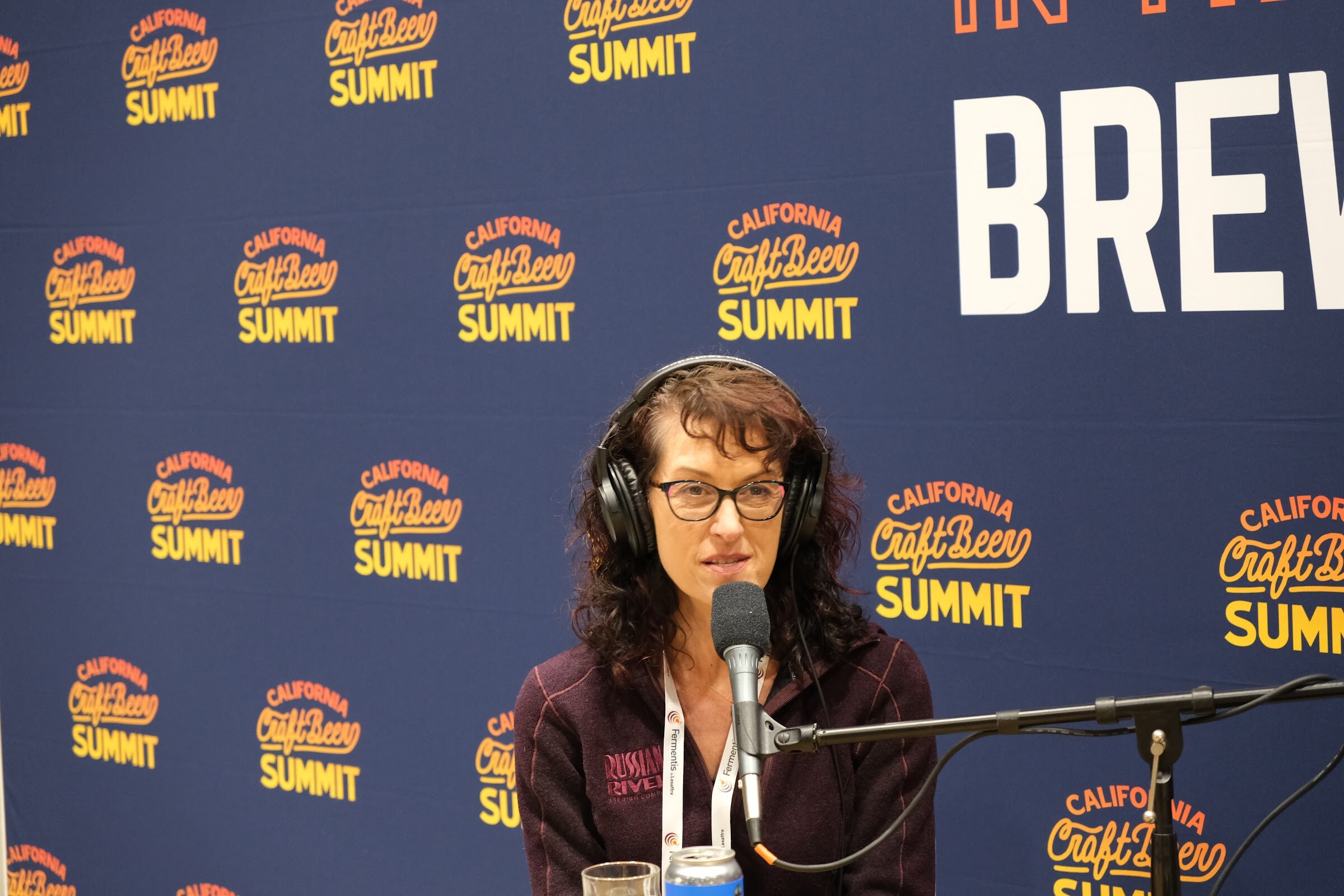 A woman in a headset speaks into a microphone in front of the California Craft Beer Summit backdrop with a beer can on the table