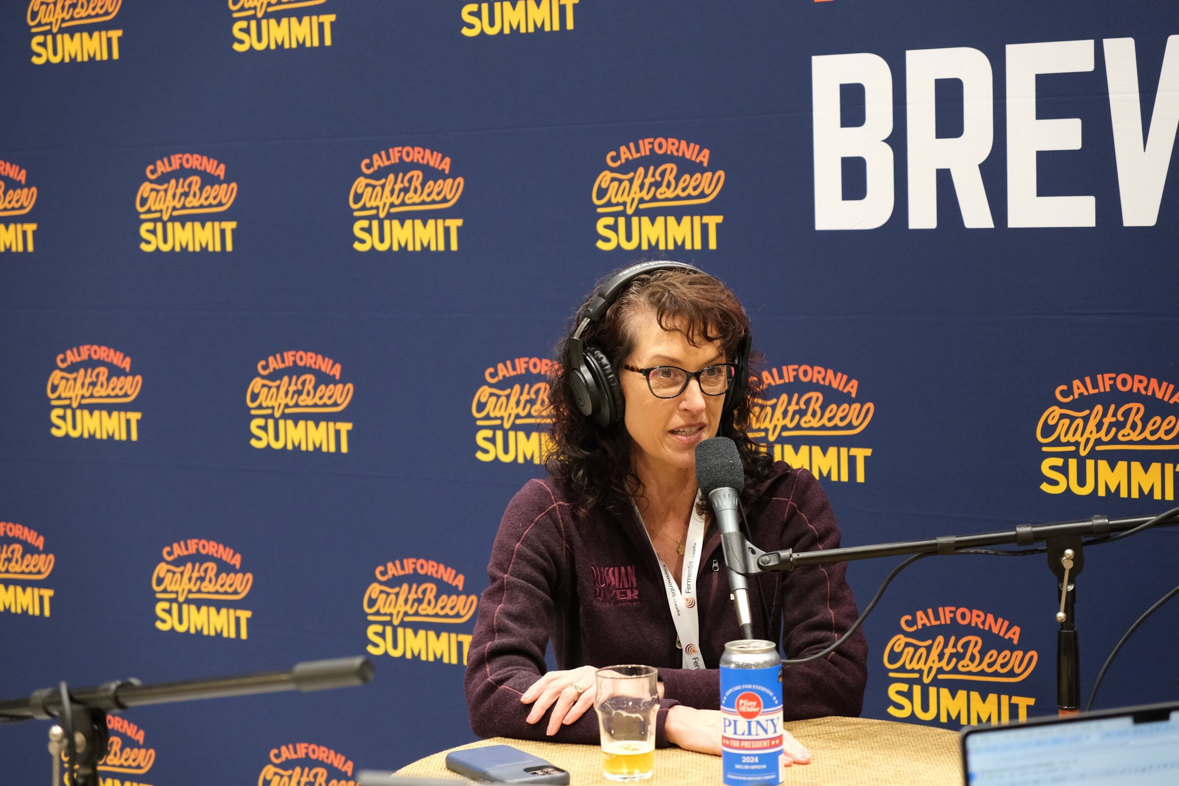 A woman in headphones speaks at the podcast table with a beer glass and a can on the table in front of a Brewbound backdrop