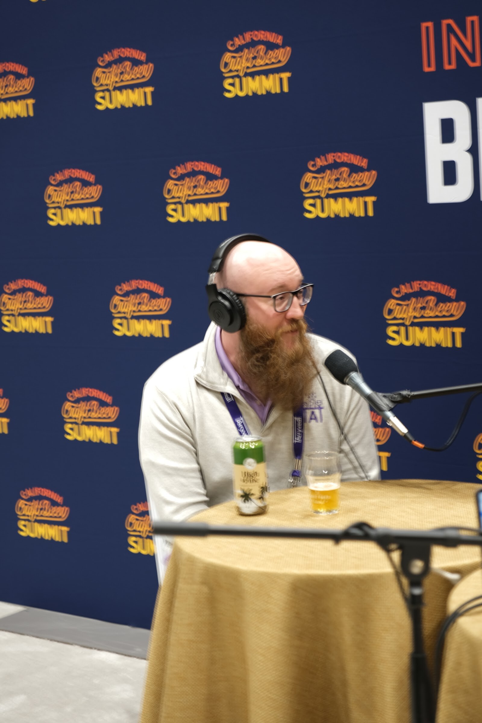 A bearded man in headphones speaks into a microphone at the podcast table with a beer beside him