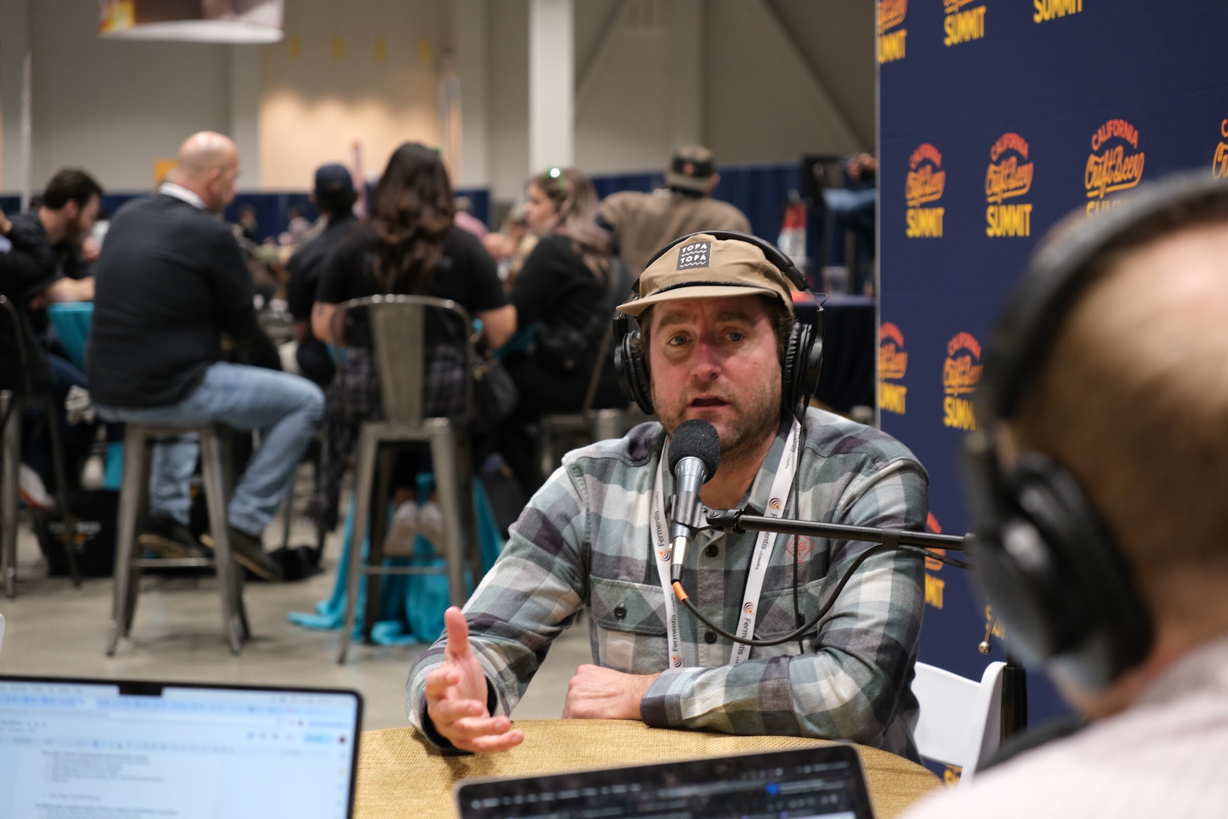 A man in a plaid flannel shirt and headphones talks into a microphone with a blurred crowd of seated attendees behind him
