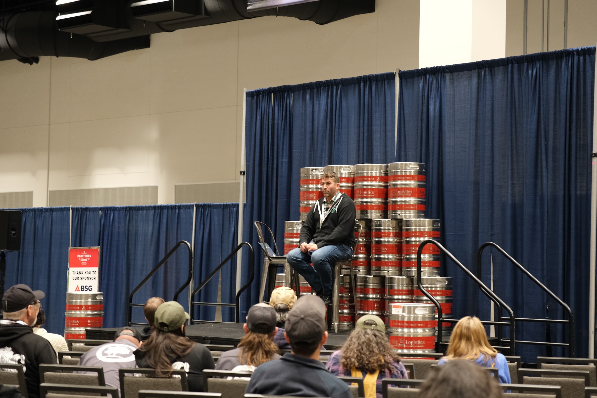 A person seated on a stool in front of a stack of kegs on a stage with an audience seated in front
