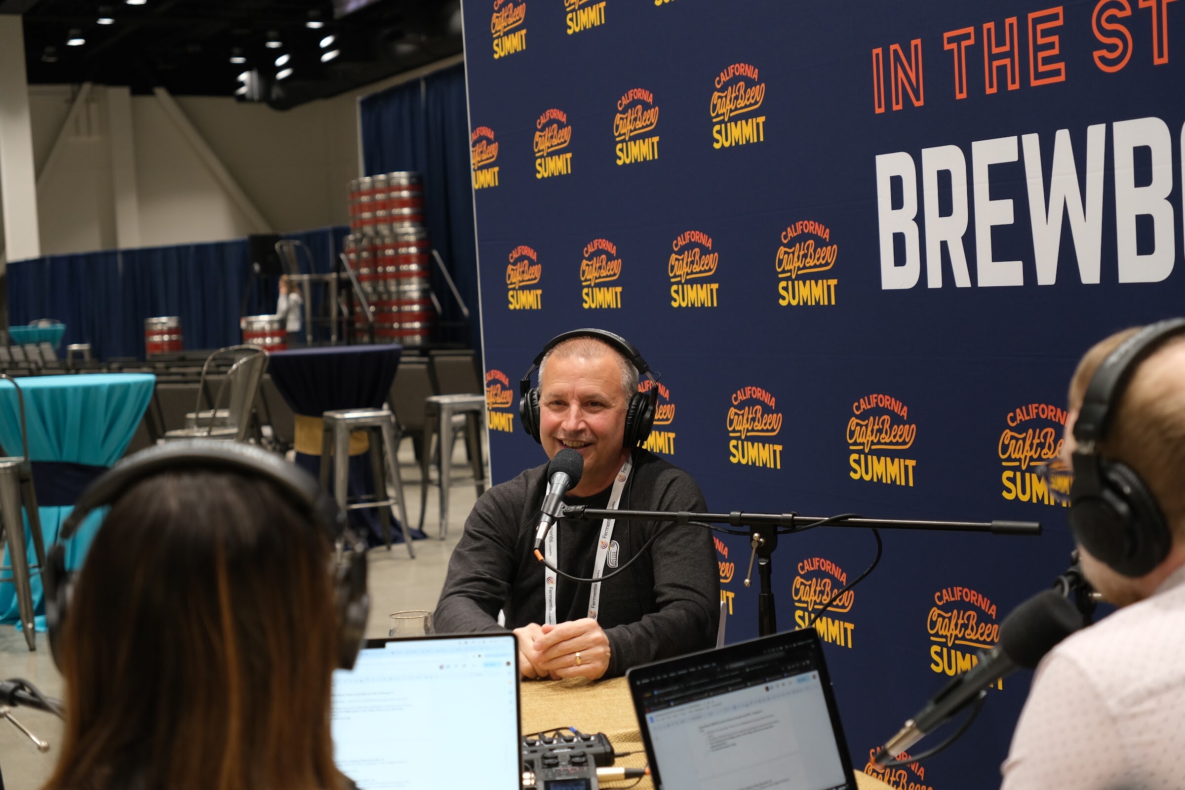 A man in a black jacket and conference lanyards gestures while speaking into a microphone in front of the Brewbound backdrop