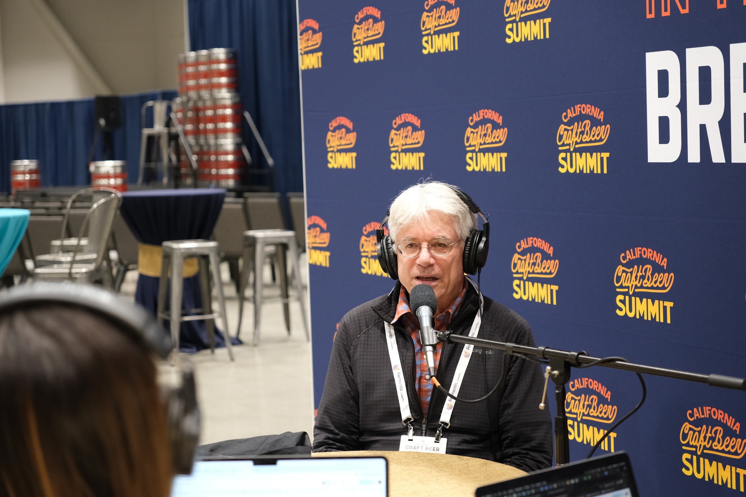A white-haired man in headphones and a lanyard speaks into a microphone in front of a California Craft Beer Summit backdrop