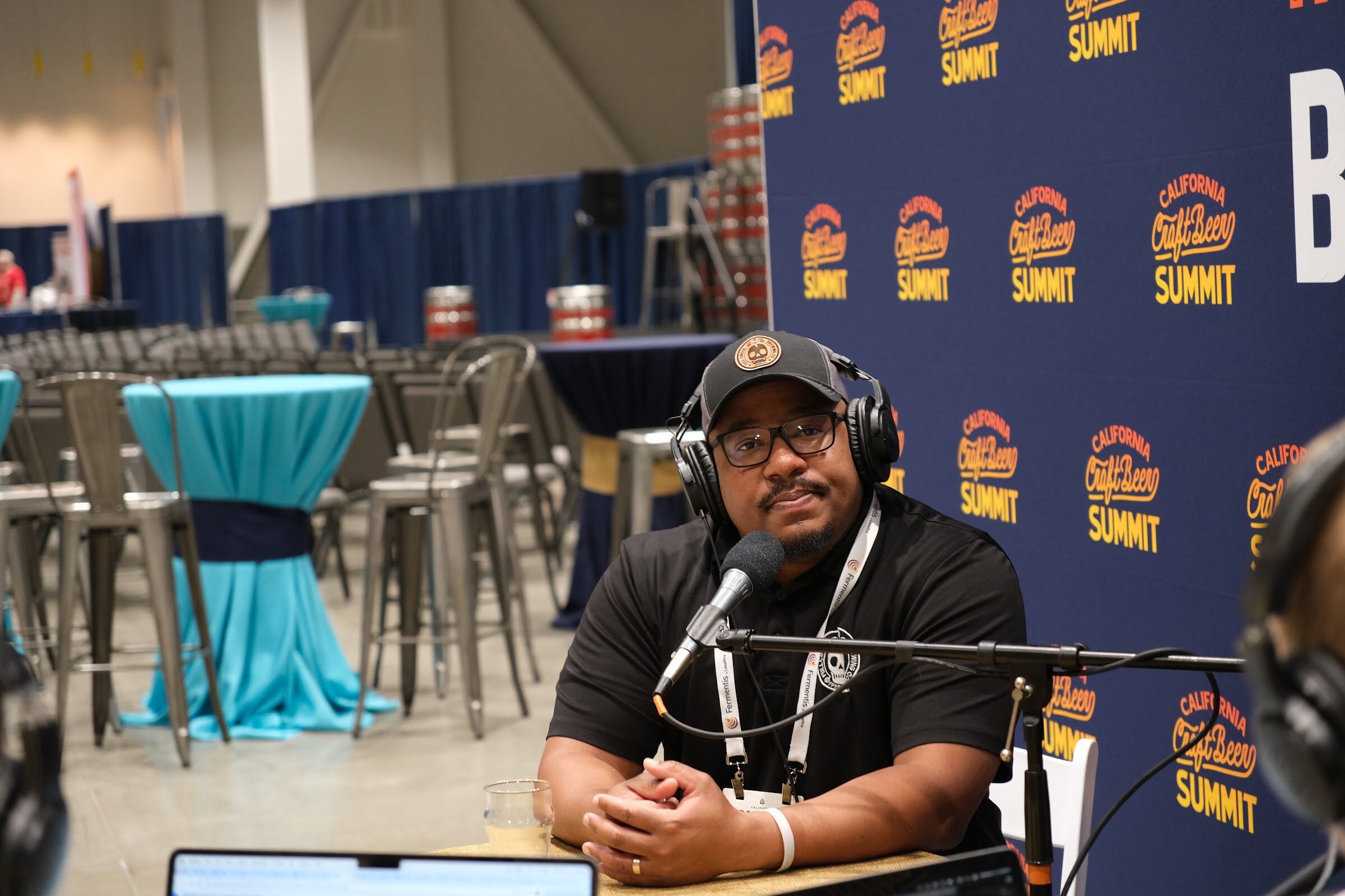 A man in a cap, headphones and a black polo smiles with hands clasped at the podcast table in front of a tabled seating area