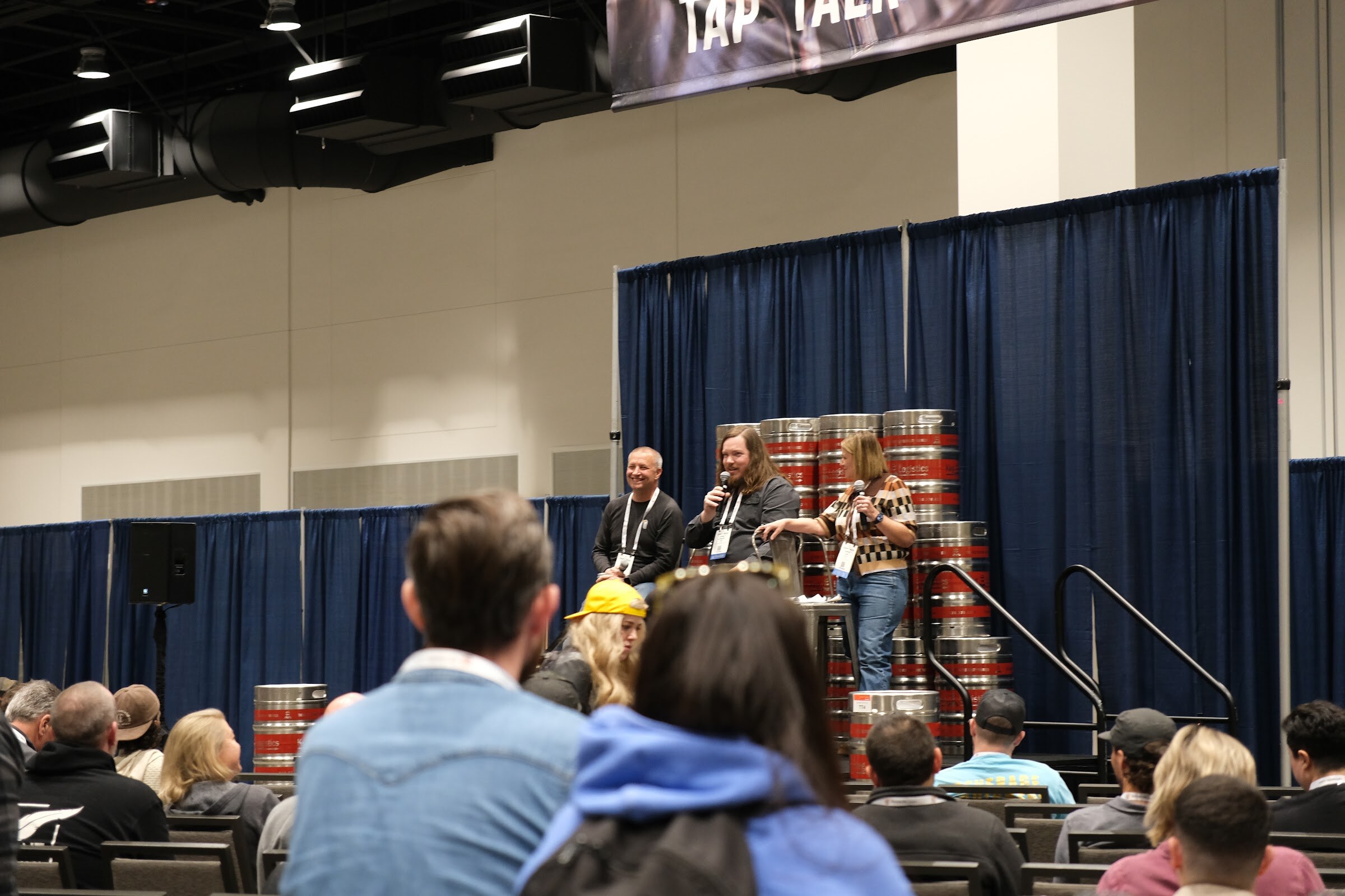 Three people seated on stools in front of a wall of kegs on a stage under a Tap Talk banner, with an audience in the foreground
