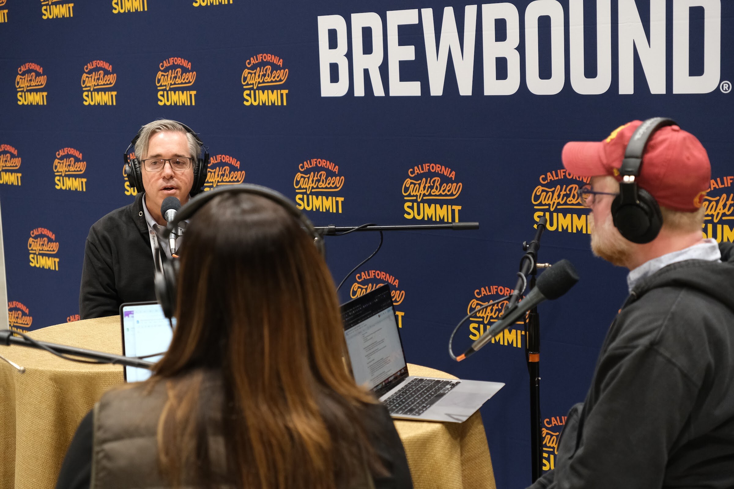 A man in a gray jacket speaks at a podcast table as a host in a red cap and another host in headphones listen