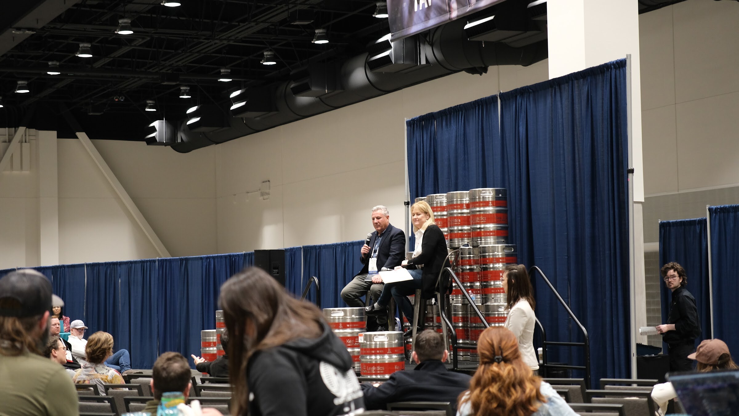 Two seated speakers talk on a stage in front of stacked kegs while attendees watch from rows of chairs