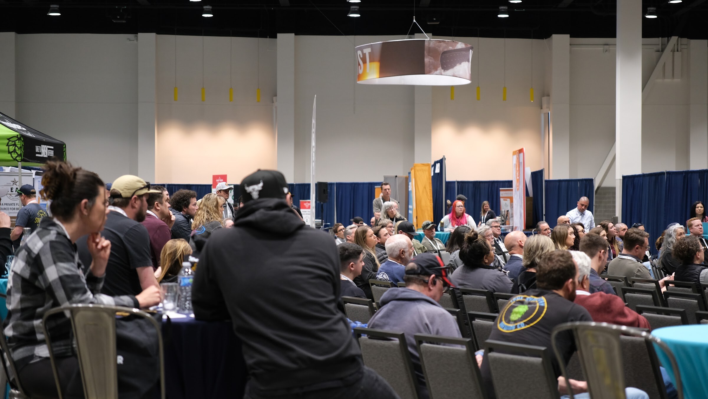 Attendees seated at tables in a large hall listening to a session, with hanging signs overhead