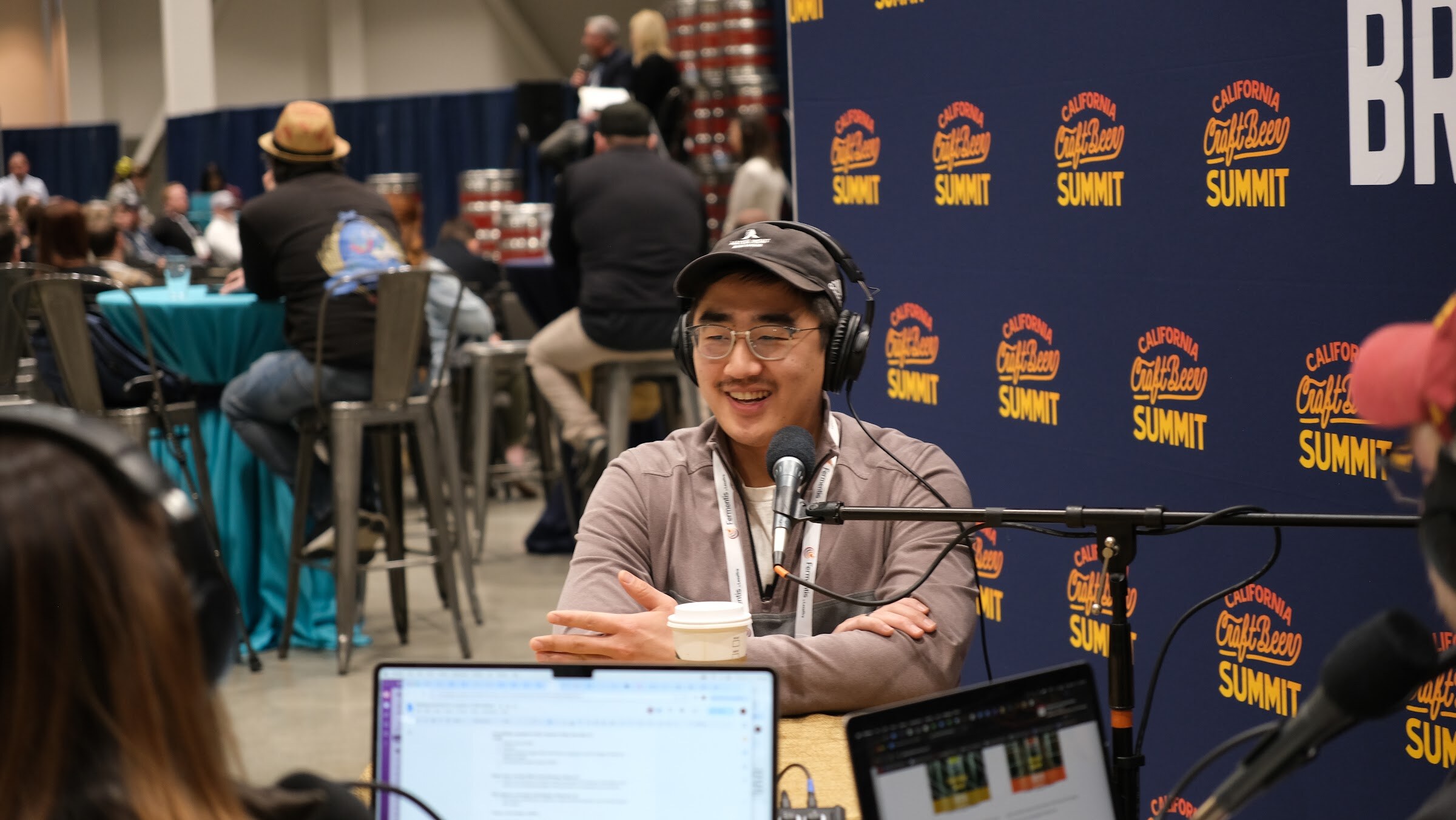 A man with glasses and a cap smiles at the podcast table with a coffee cup in front of a Brewbound backdrop and a blurred room