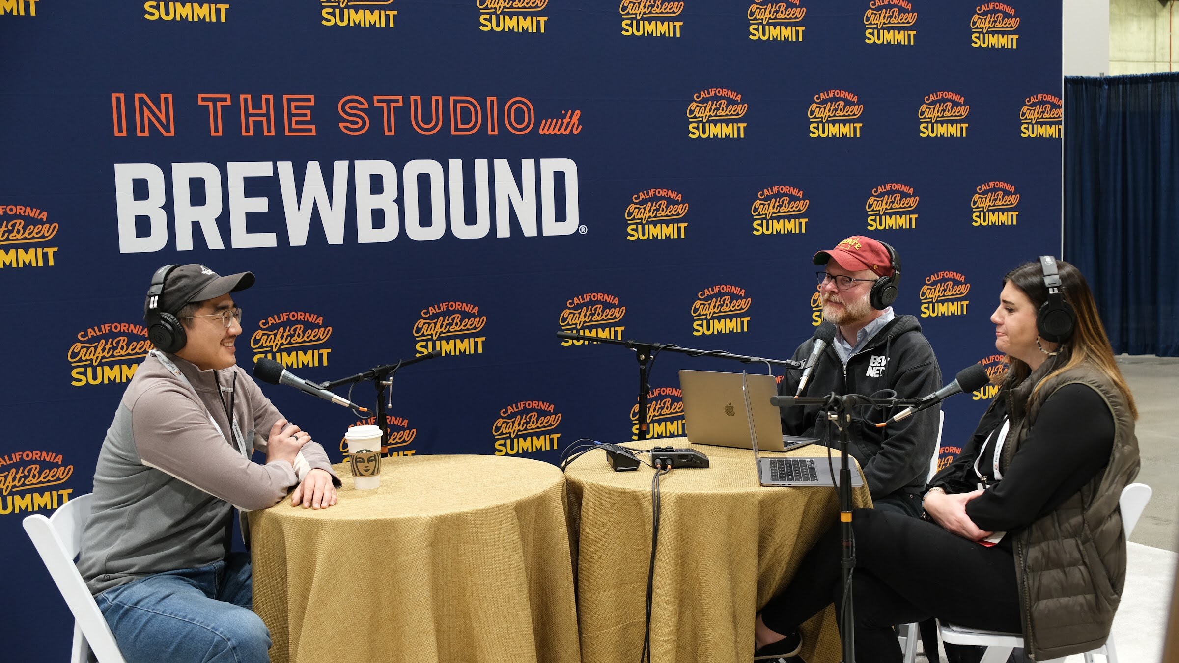 A guest in a gray jacket sits across a round table from two hosts in headphones in front of the In the Studio with Brewbound backdrop