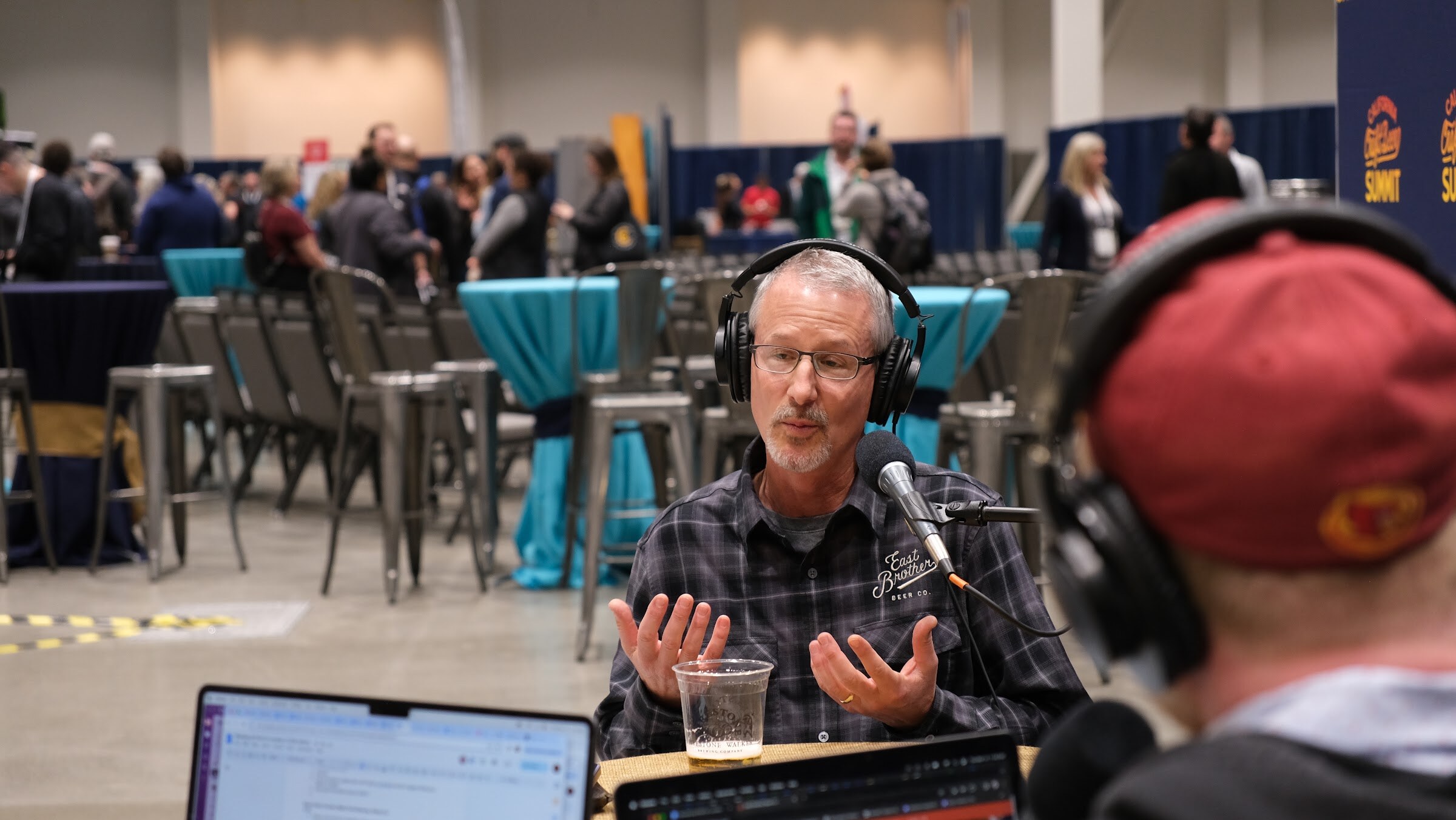 A man in a plaid shirt and headphones gestures with open hands while a host in a red cap listens in the foreground
