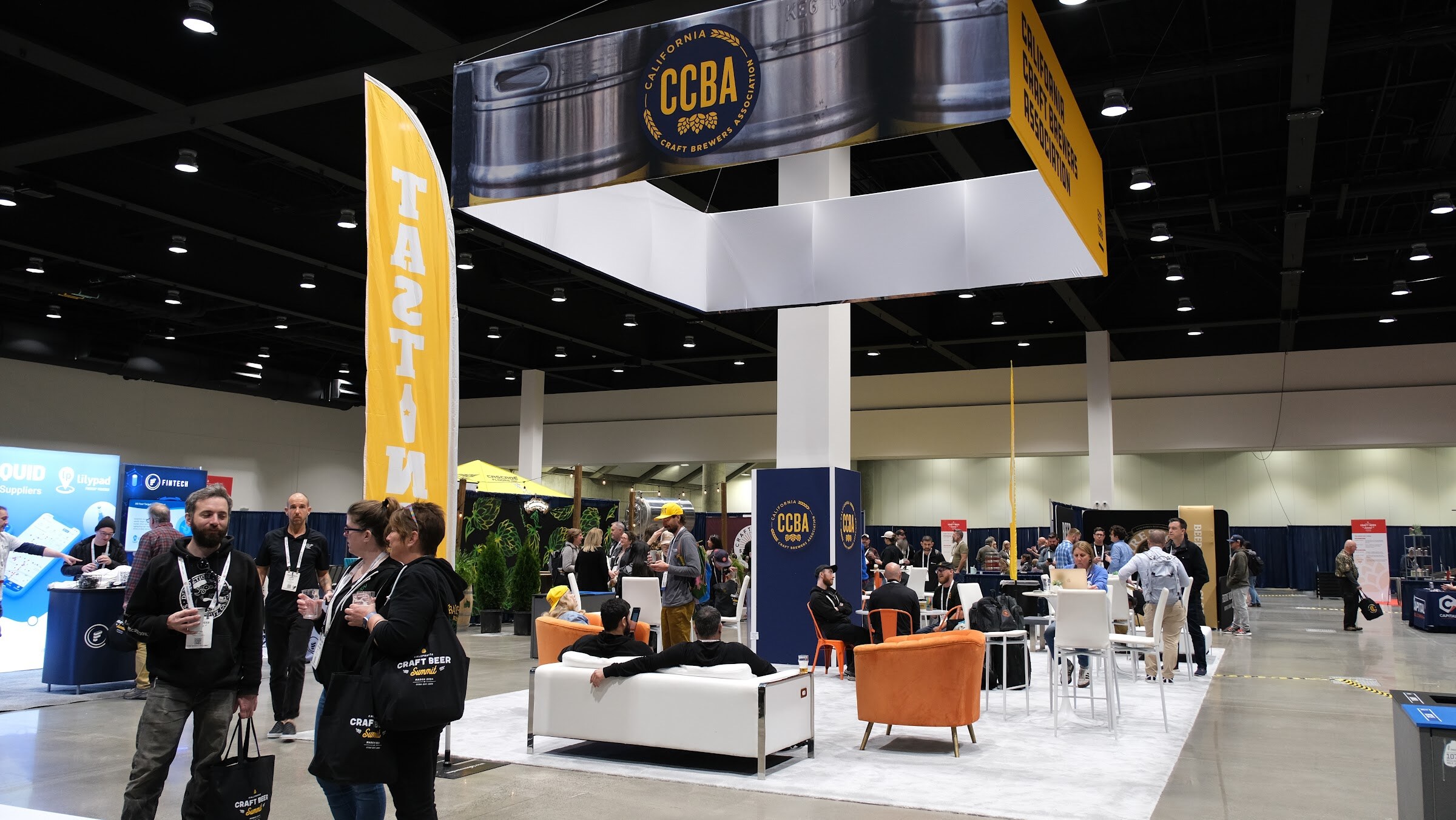 A lounge-style booth with white sofas and orange chairs under a hanging California Craft Brewers Association sign on the expo floor