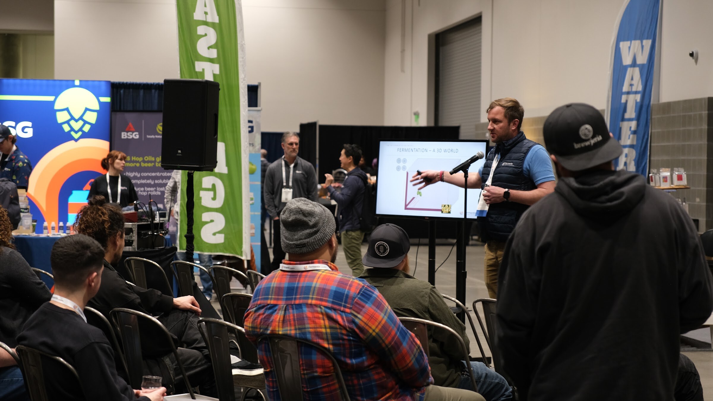A speaker presents beside a screen with a chart to an audience seated on the expo floor, with banners in the background