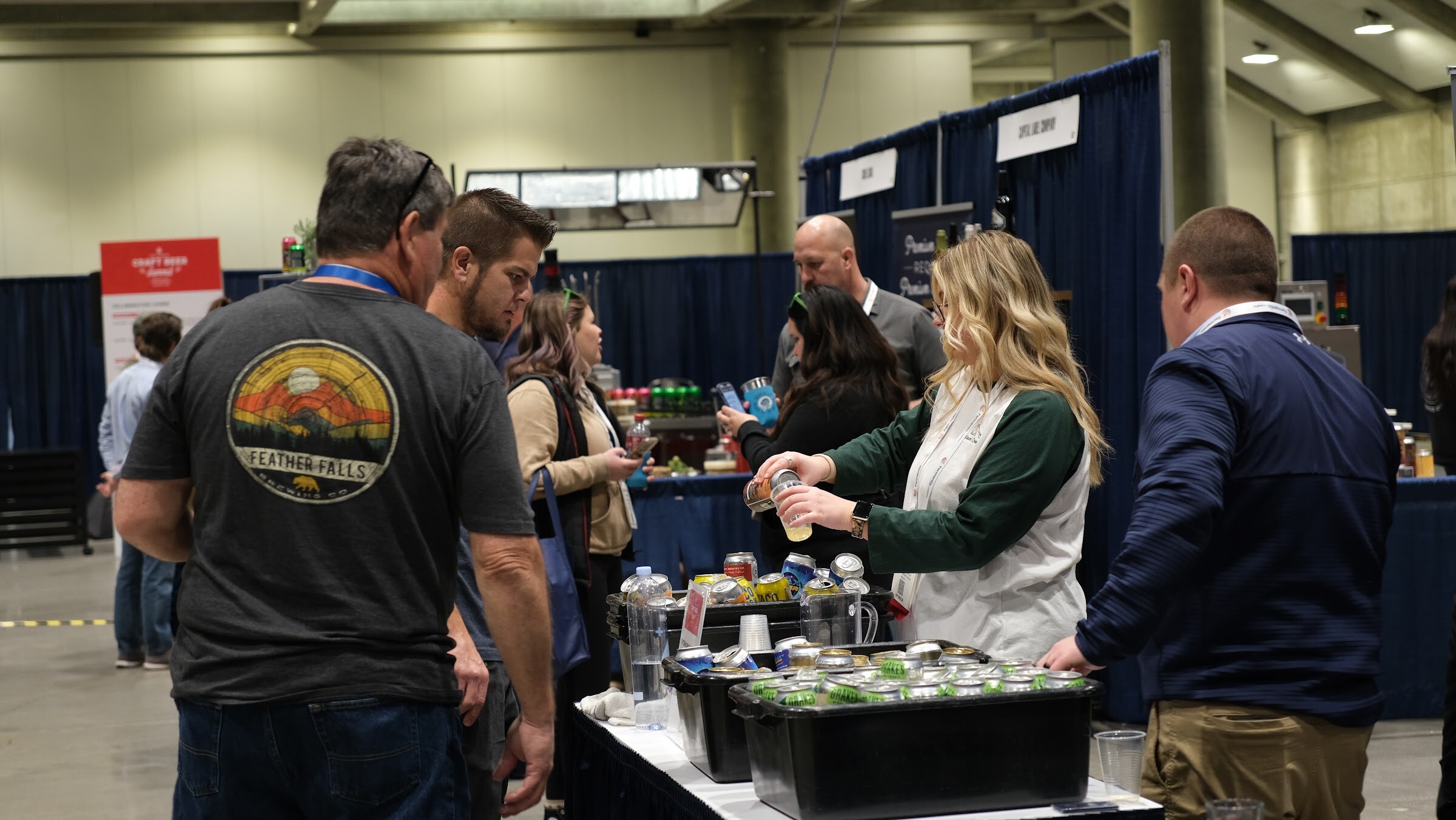 Attendees gather around a table of beer and supplies while a woman in a white vest pours samples