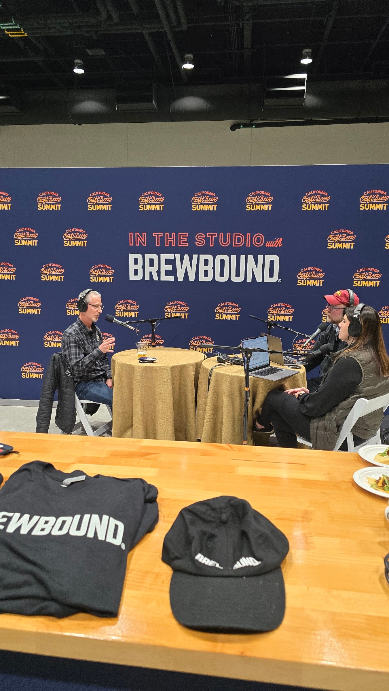 A wooden table with Brewbound T-shirts and a black Brewbound cap in front of the podcast studio backdrop