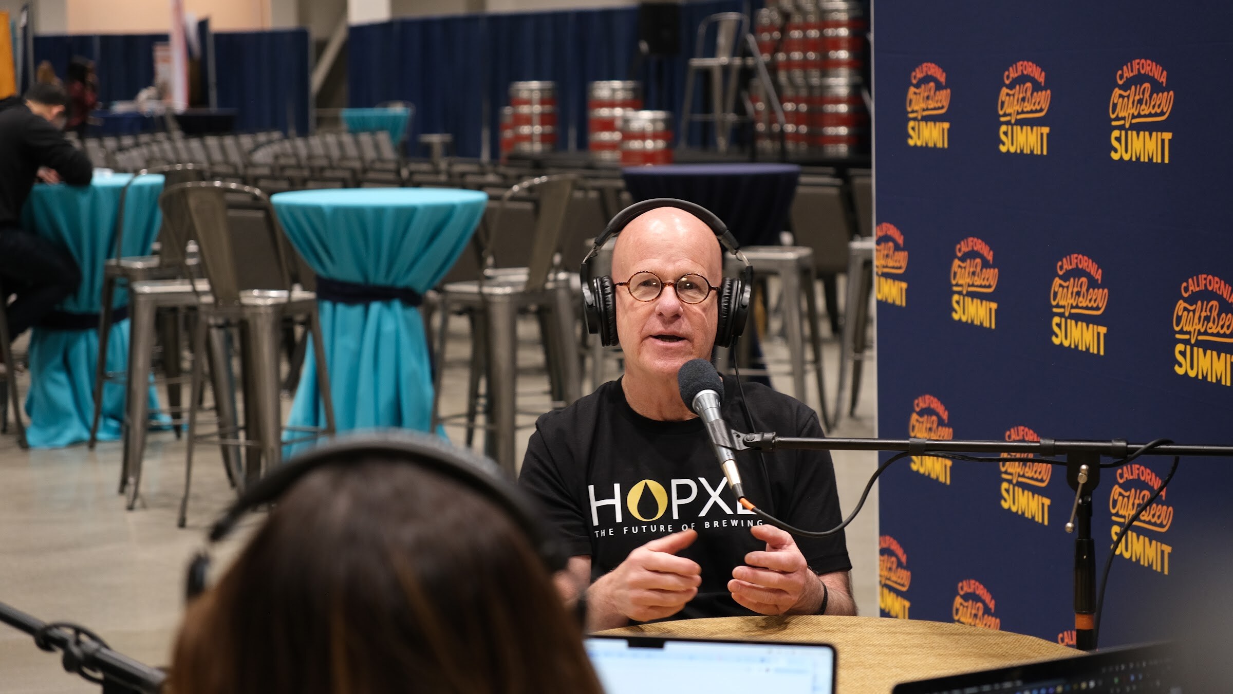 A bald man in a HOPXL shirt gestures while speaking into a microphone at the podcast table
