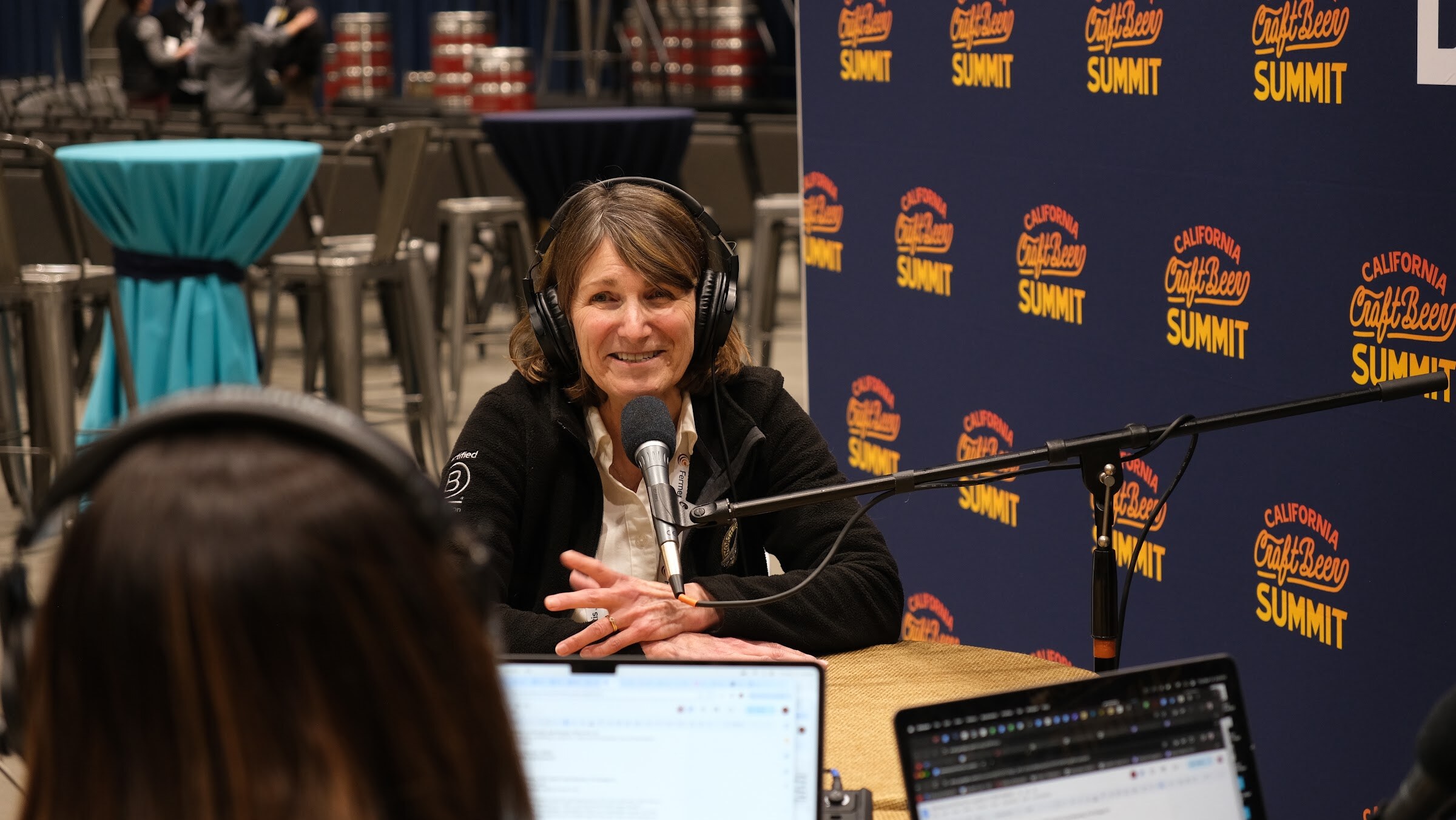 A woman with long hair in a black jacket and headphones smiles at the podcast table in front of the Brewbound backdrop