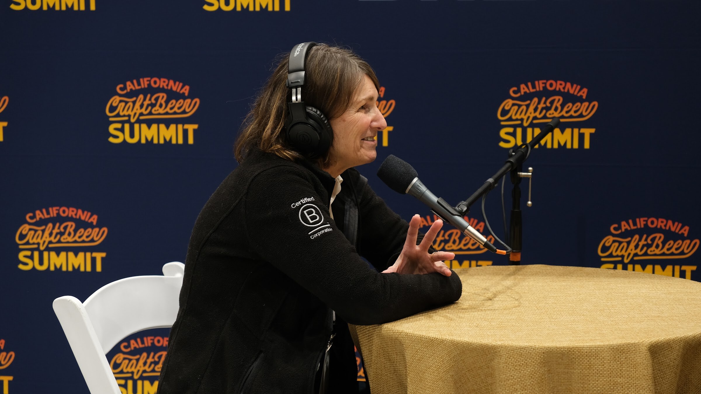 A woman in a black jacket and headphones speaks into a microphone seated at a round table with a gold cloth