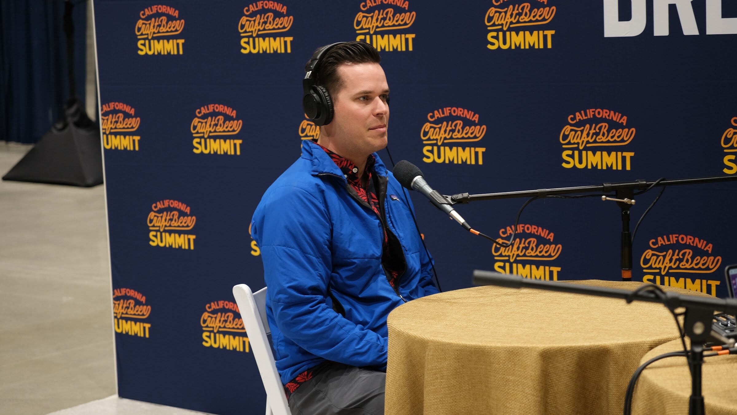 A man in a blue jacket and headphones sits at a podcast table with a microphone in front of the California Craft Beer Summit backdrop