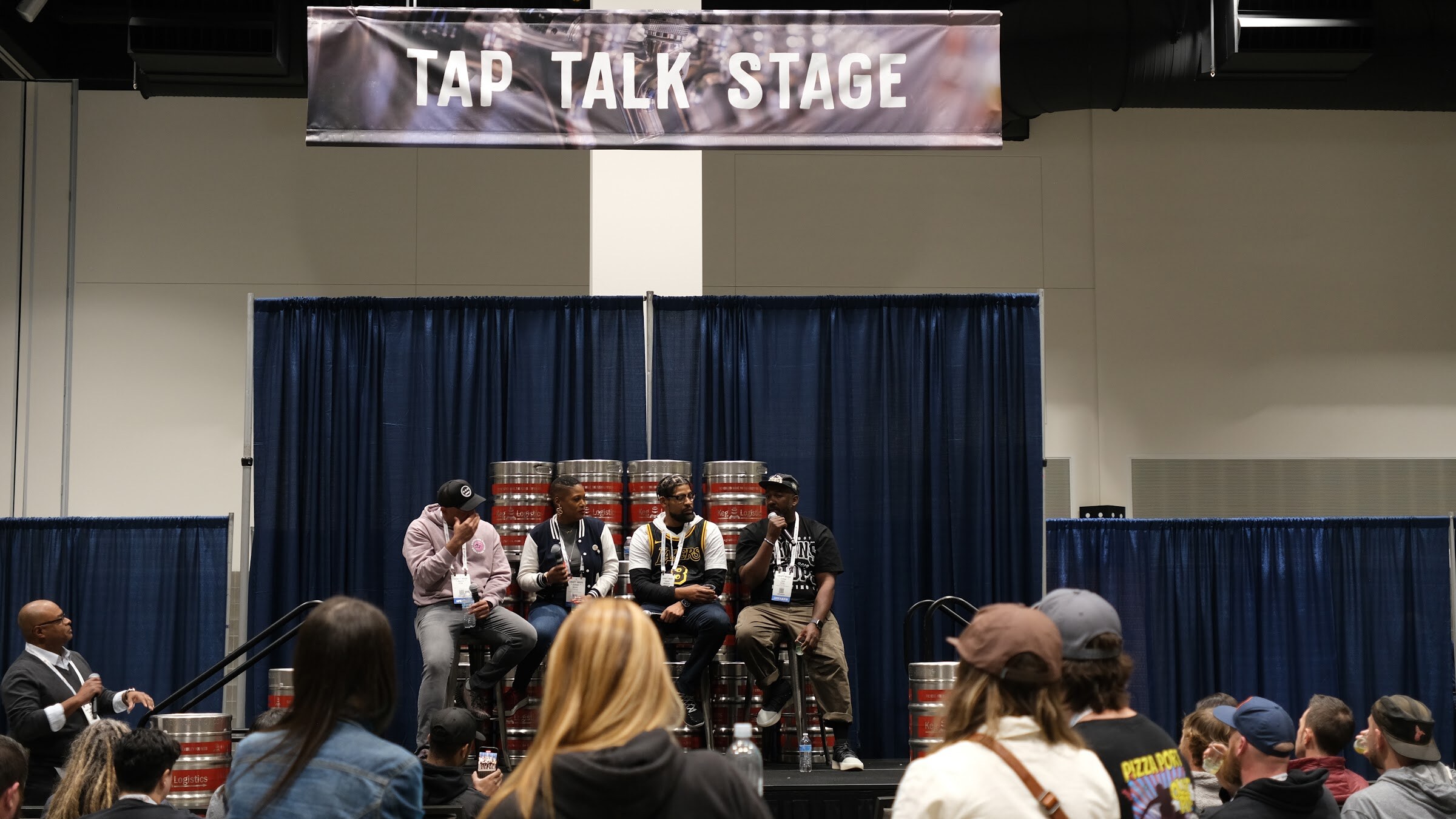 Five panelists seated on stools in front of stacked kegs under a Tap Talk Stage banner with attendees watching
