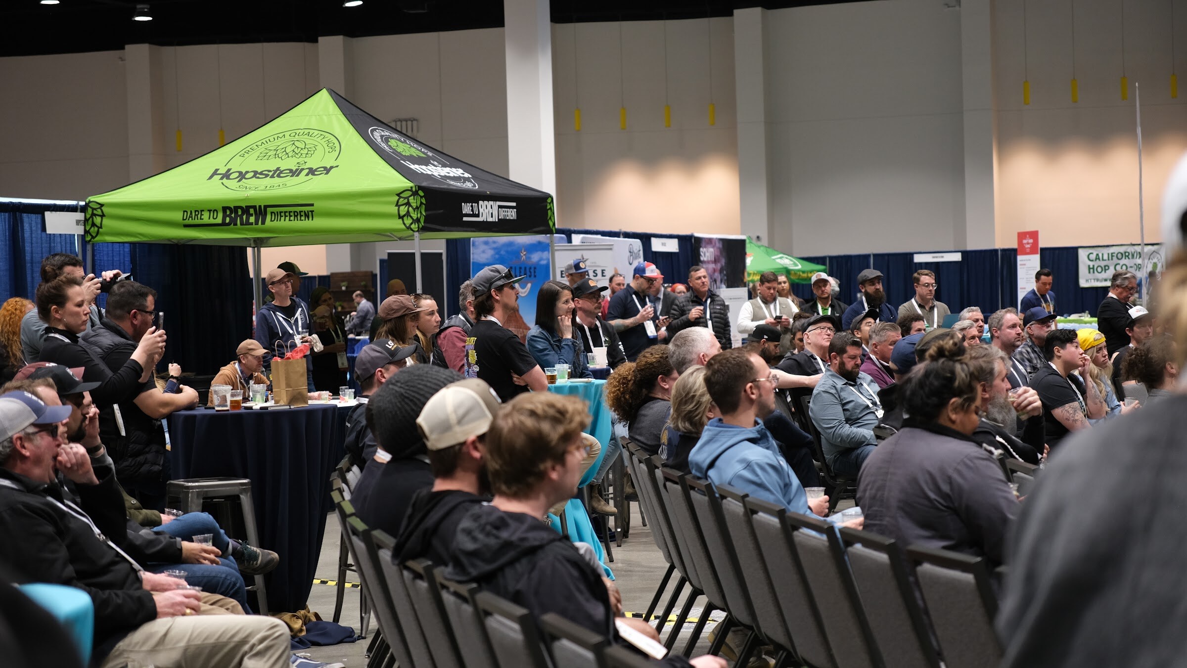 Rows of attendees seated in a hall watching a session, with a green Hopsteiner tent in the background