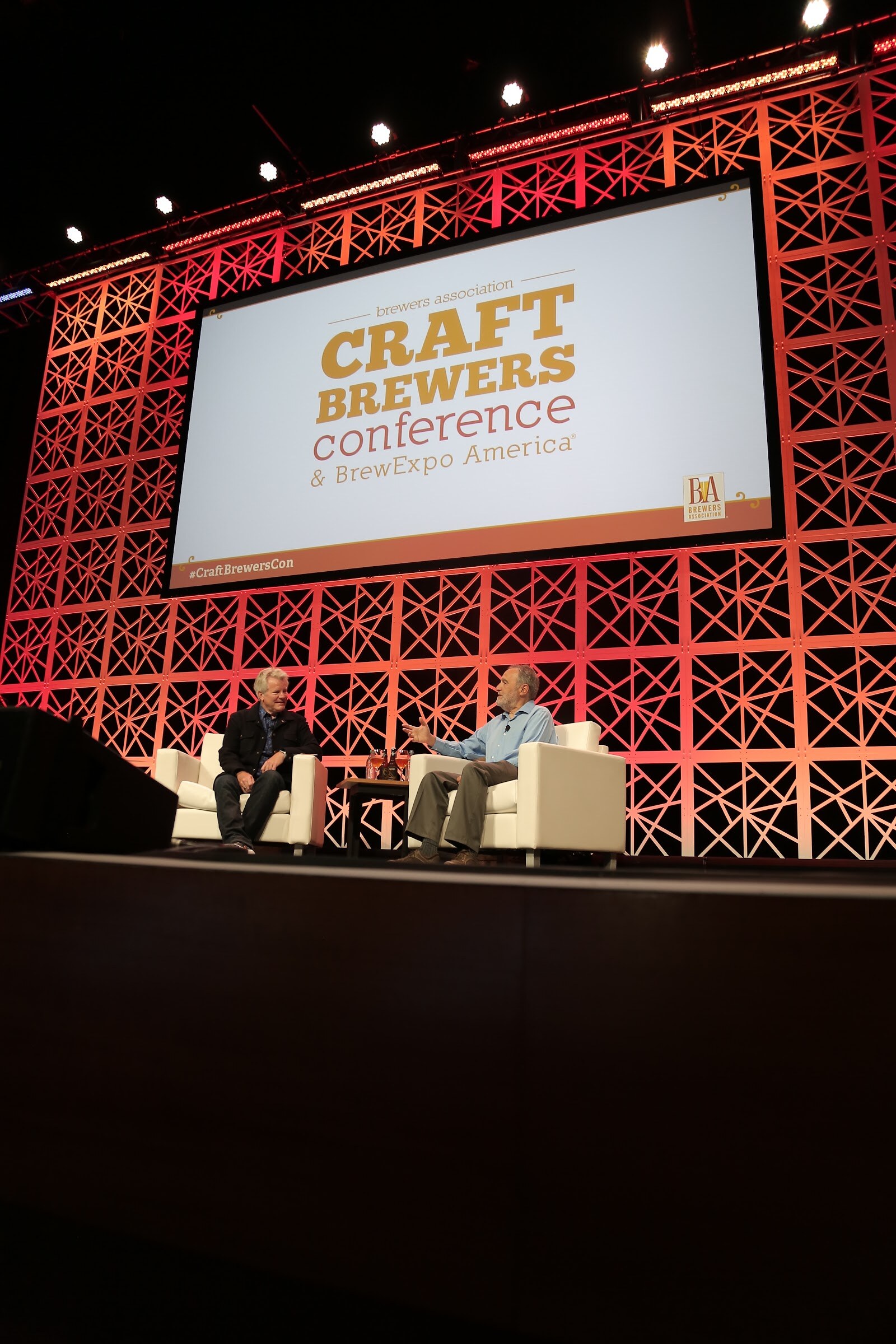 Vertical wide shot of two people seated in white armchairs on a stage in front of a red lattice backdrop and a large screen reading Craft Brewers Conference & BrewExpo America.