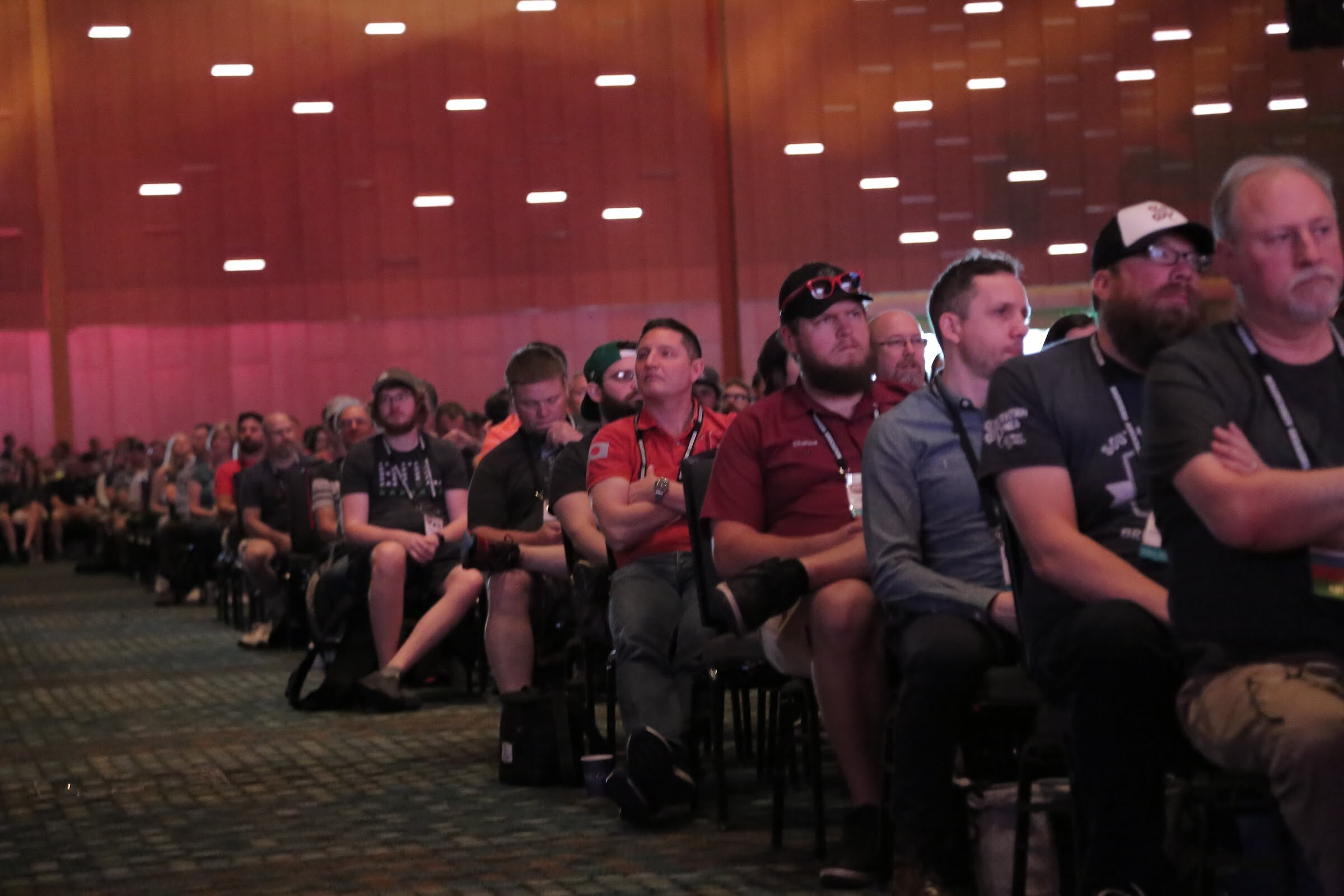 A row of badge-wearing attendees in a large ballroom, several with arms crossed, looking toward the stage.