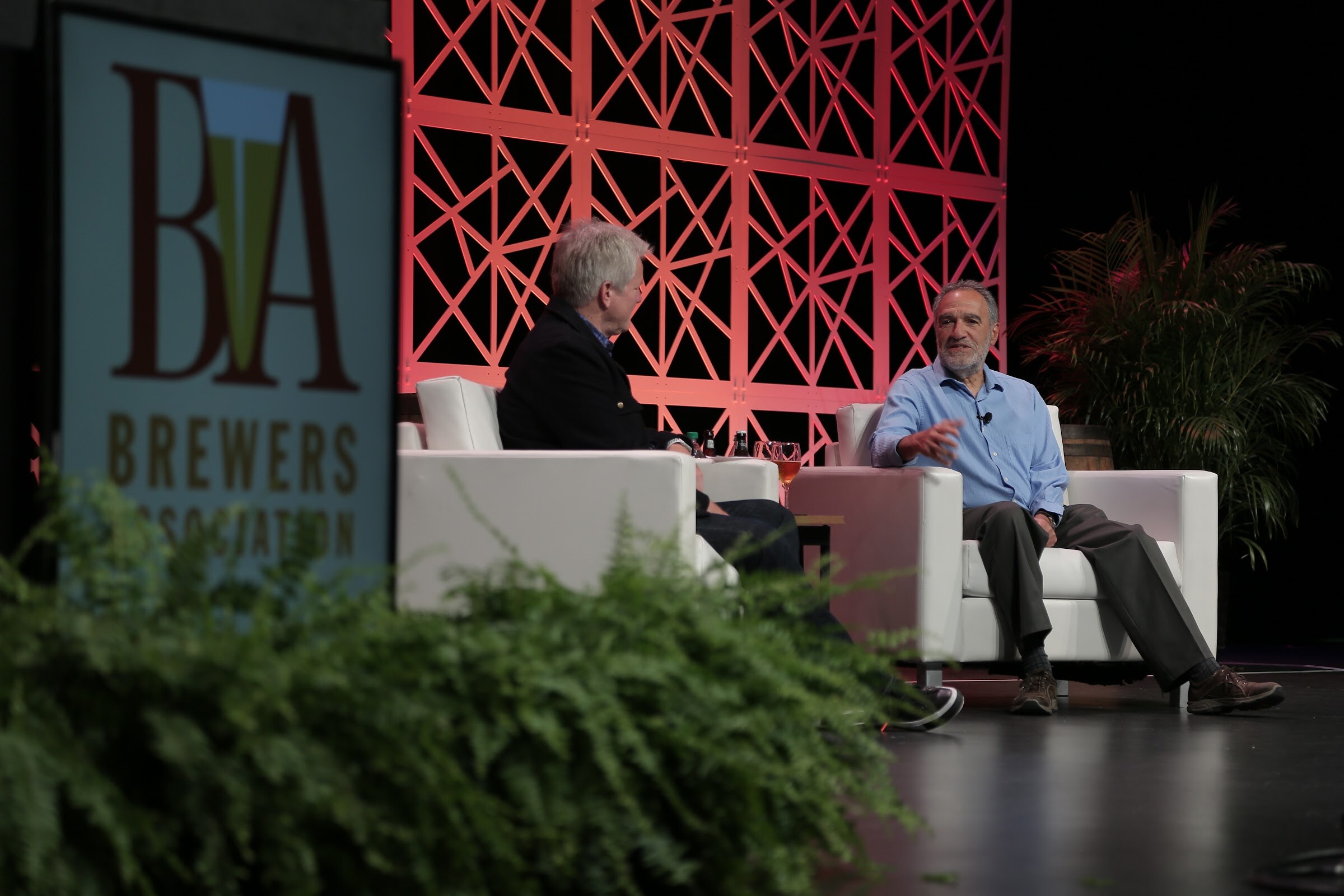 Two men in white armchairs on a stage with a red lattice wall, a Brewers Association sign at left and green ferns in the foreground.