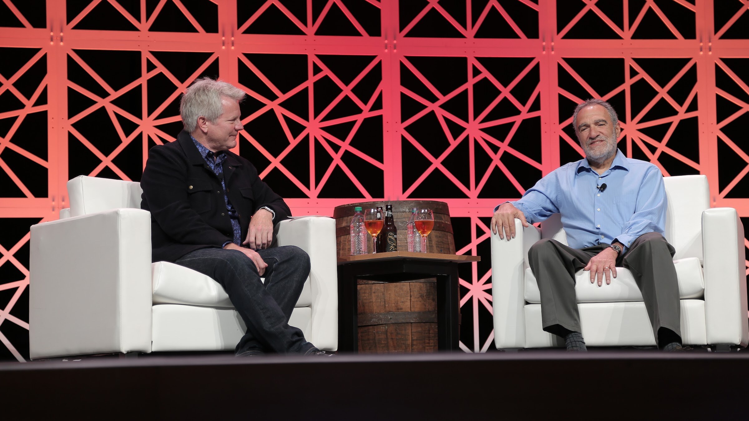 Two men sit in white armchairs on either side of a barrel table holding beer glasses, in front of a red lattice backdrop.