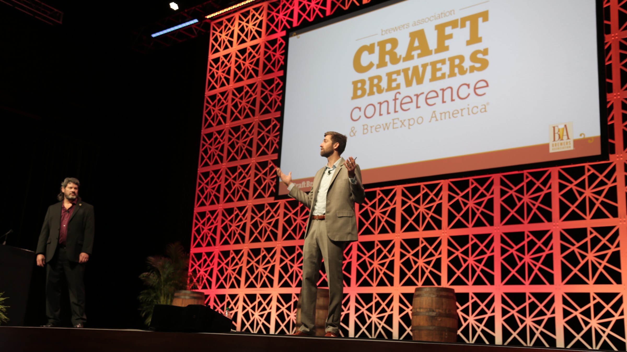 A man in a tan suit gestures with open hands on a stage under a Craft Brewers Conference & BrewExpo America screen; a second man stands at the left edge.