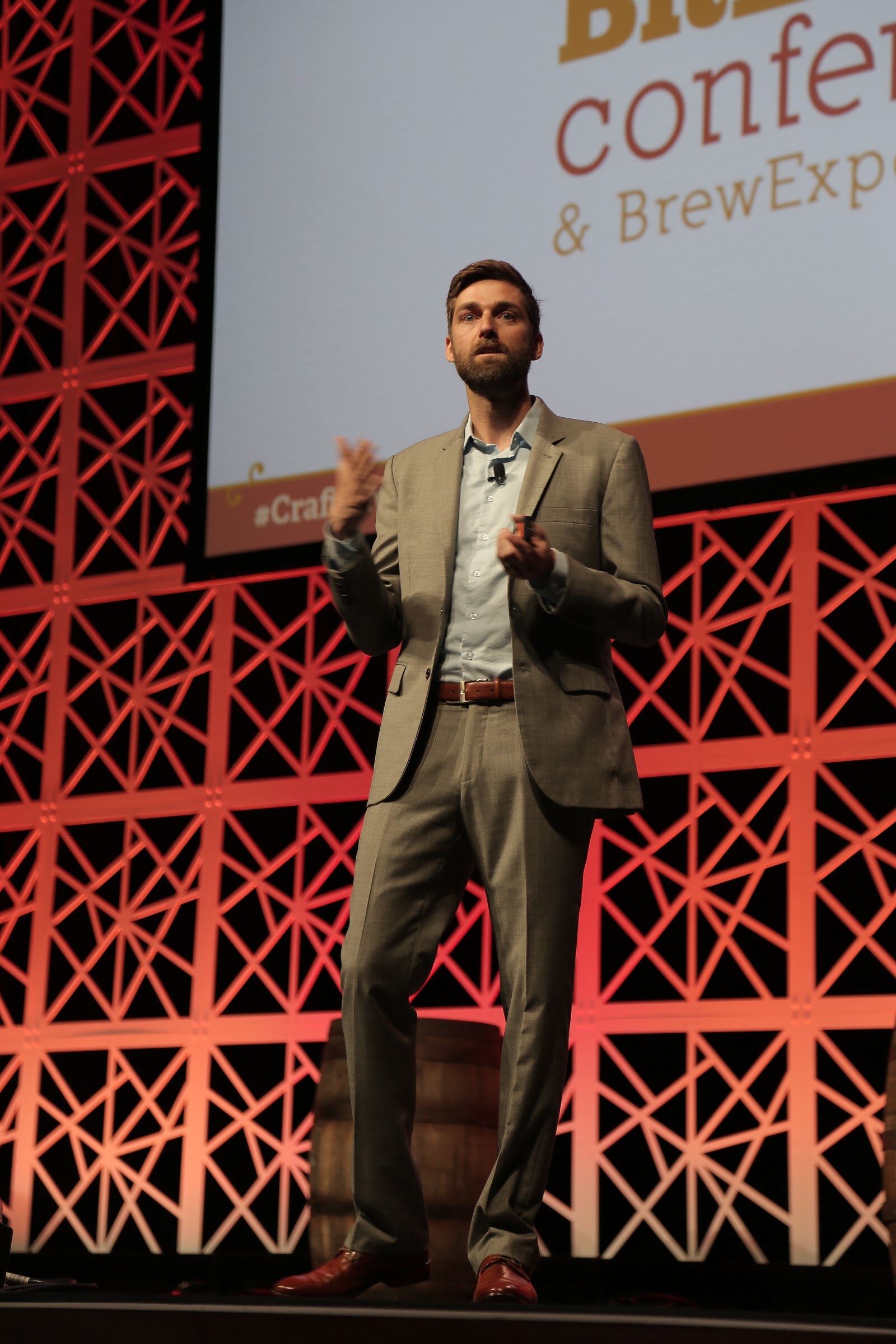 Full-length portrait of a bearded man in a tan suit and brown shoes gesturing on a stage in front of a red lattice wall.