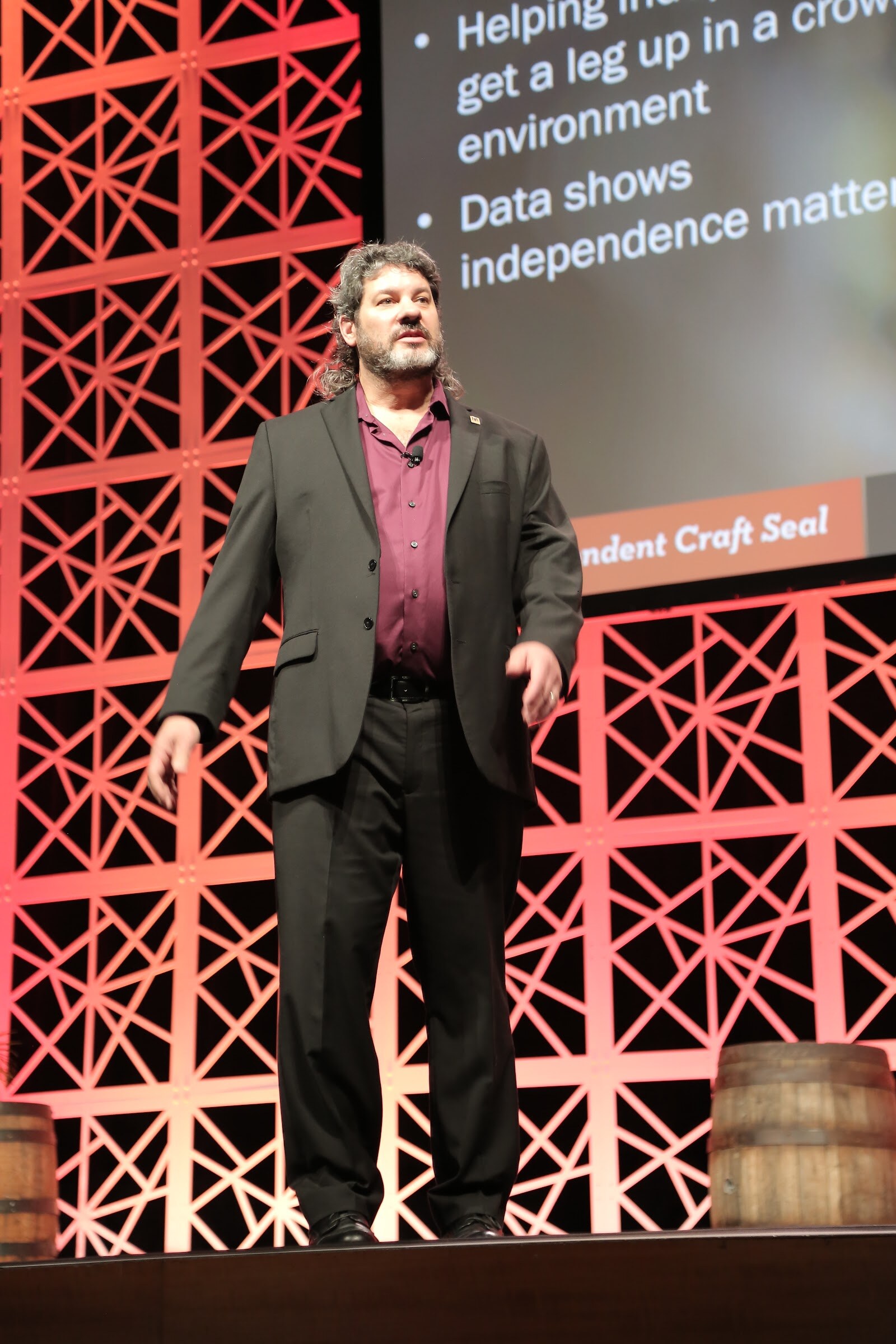A man in a purple shirt and black blazer stands on a stage in front of a screen with bullet points and the words Independent Craft Seal.