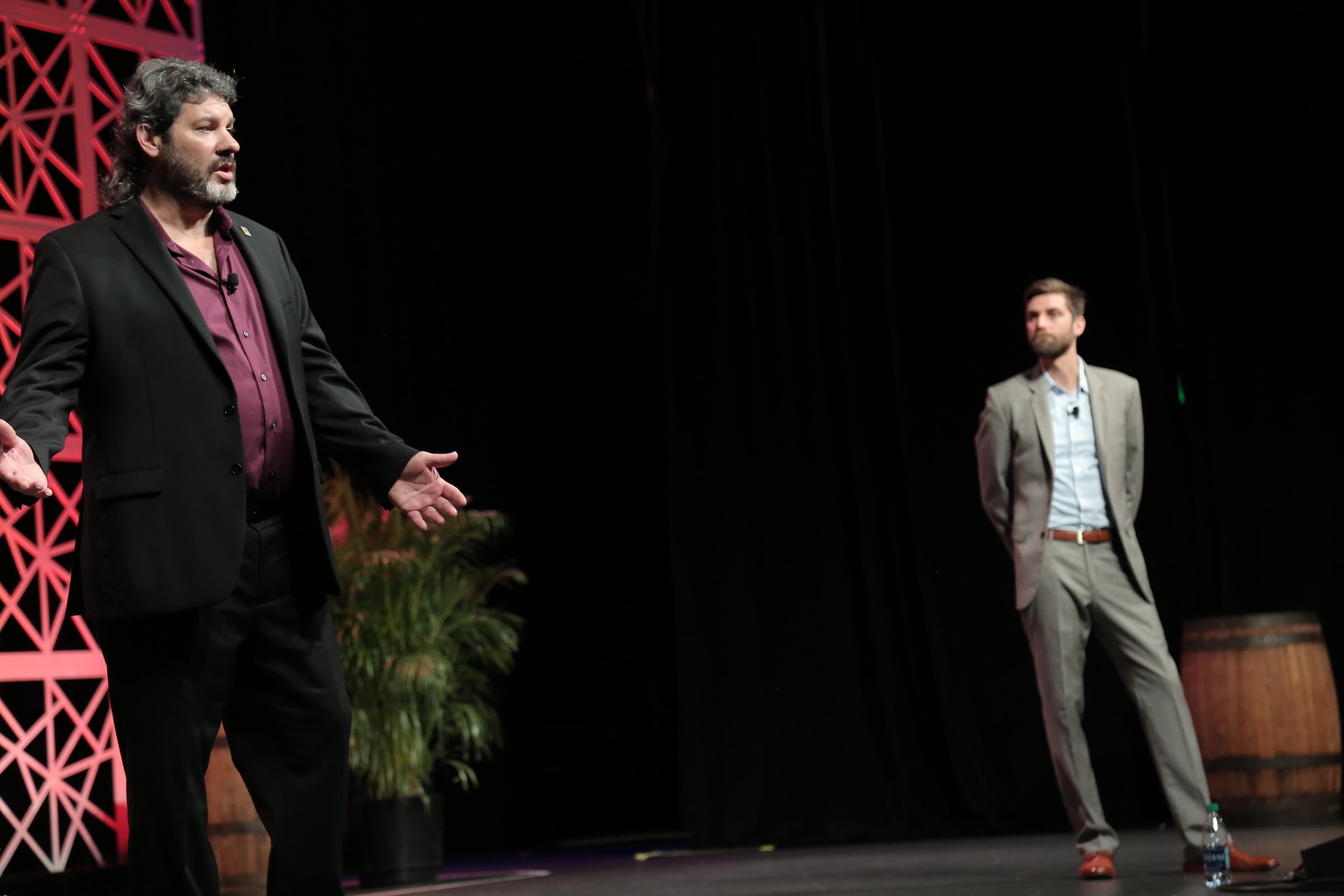 A man in a purple shirt and black blazer holds out his hands at left while a man in a gray suit stands at right beside a plant and a whiskey barrel.