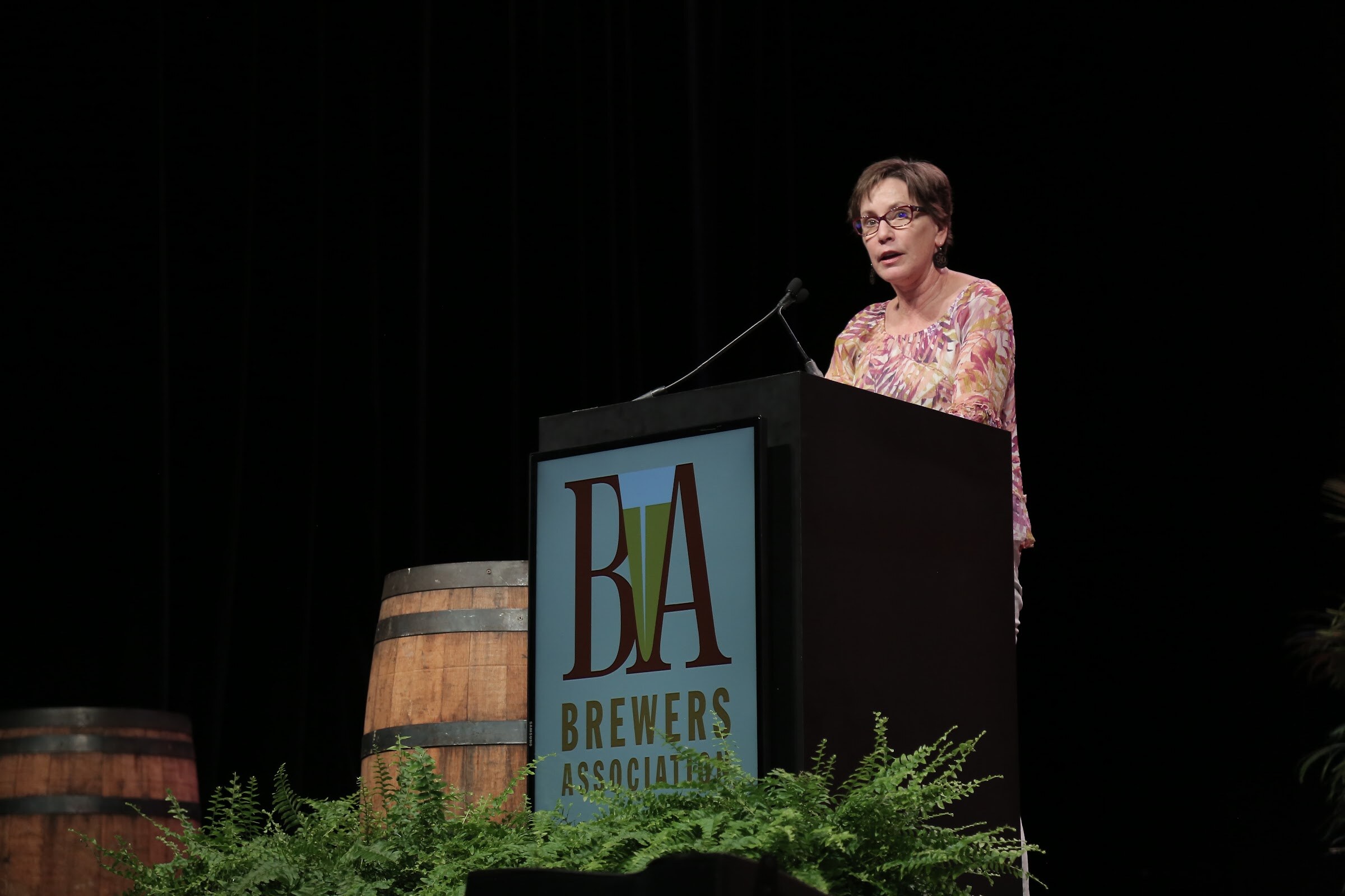 A woman with glasses speaks at a black lectern bearing a Brewers Association logo, with whiskey barrels and ferns beside it.