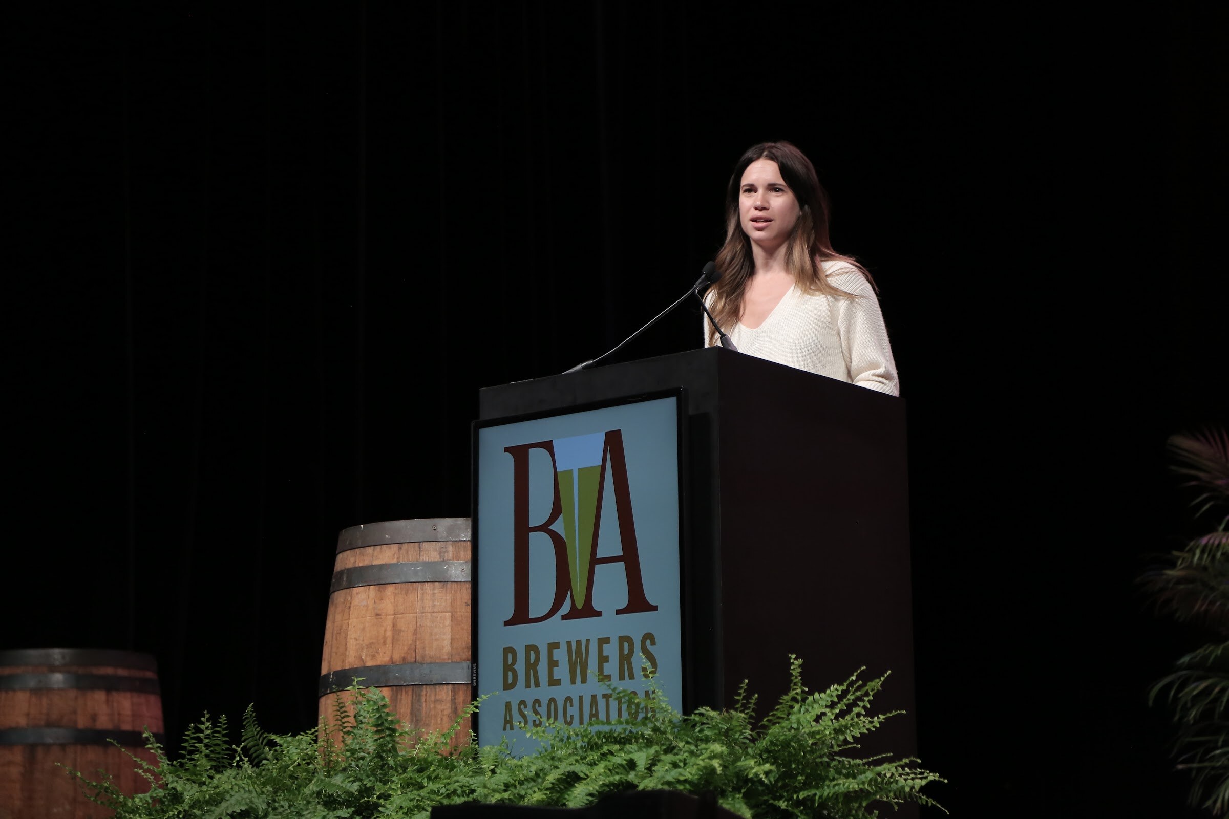 A woman with long brown hair in a cream sweater speaks into a microphone at a lectern with a Brewers Association logo, flanked by barrels and ferns.