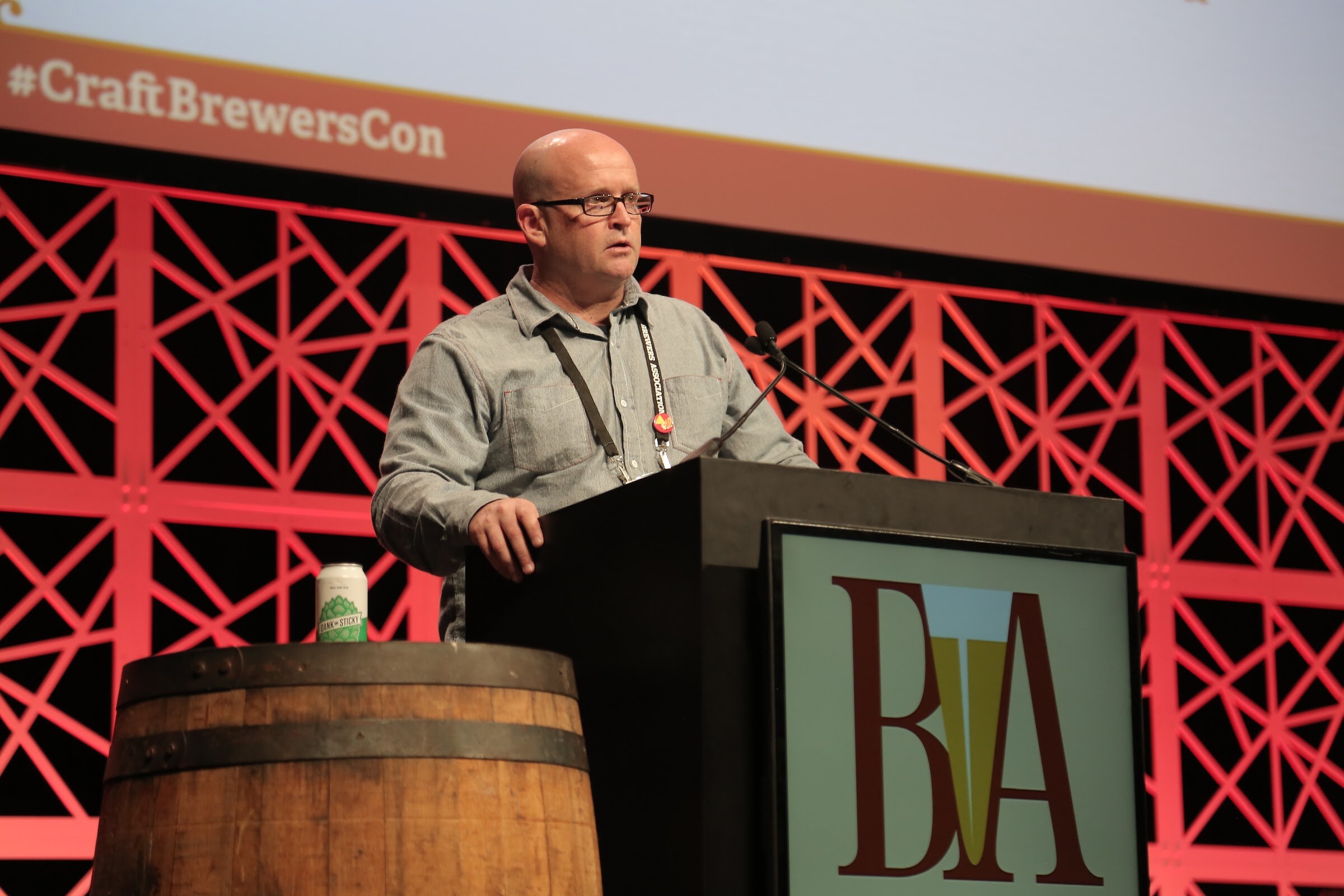 A bald man in glasses and a gray shirt speaks at a lectern marked with a BA logo, with a beer can on a barrel and the text #CraftBrewersCon behind him.