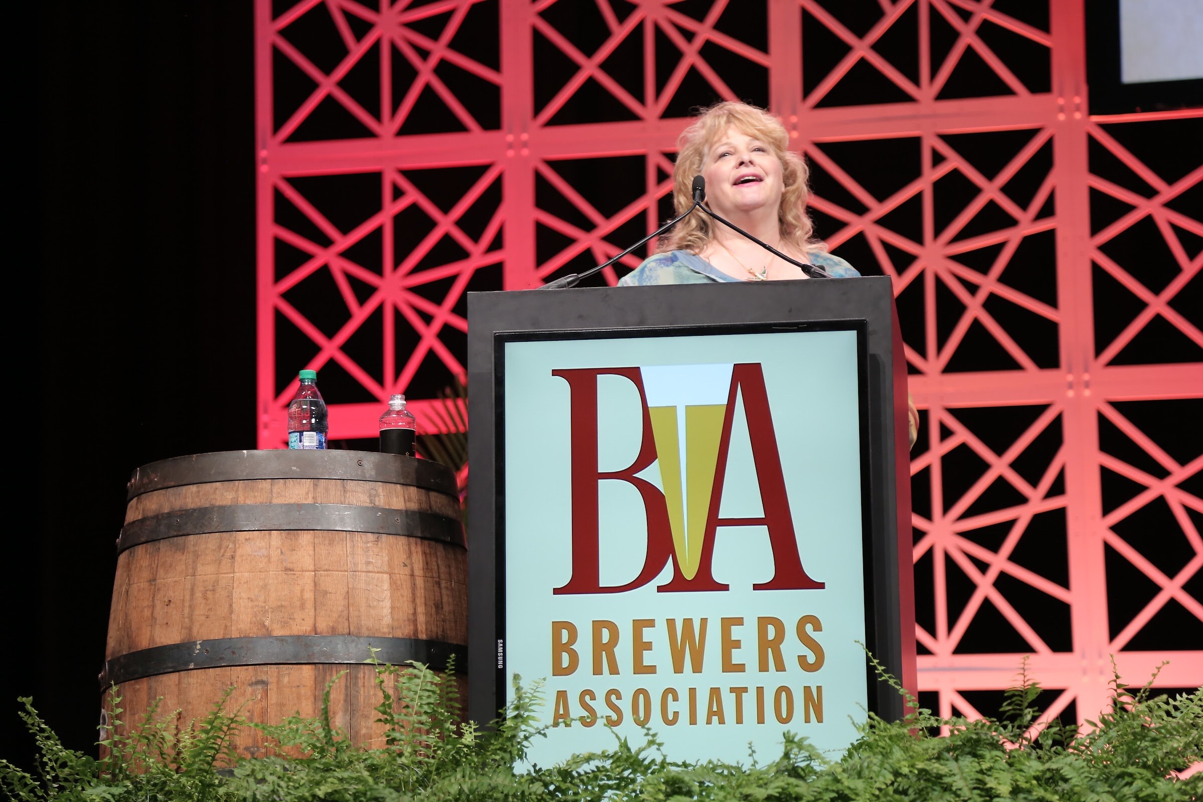 A woman with curly blonde hair speaks at a lectern with a large Brewers Association logo, with a barrel and ferns in the foreground.
