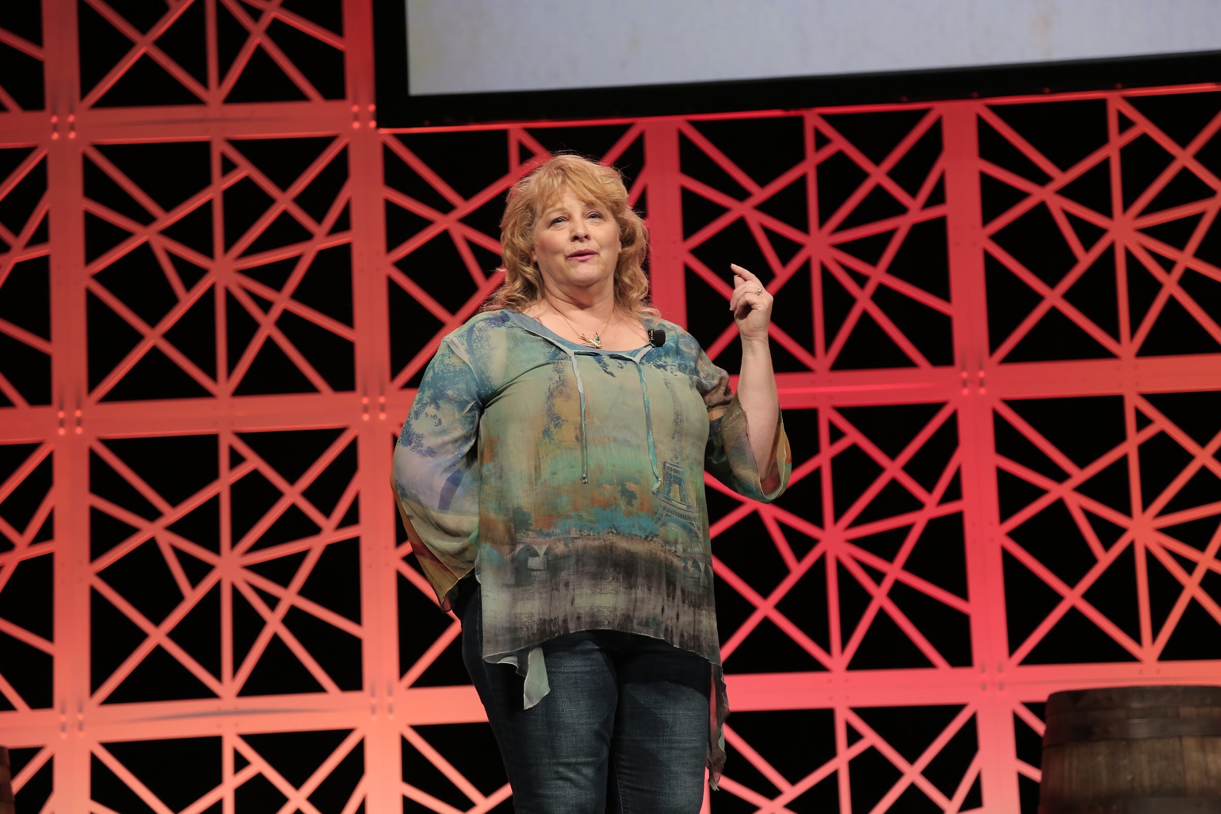 A woman in a blue-green patterned blouse and jeans stands in front of a red lattice backdrop with one hand raised.