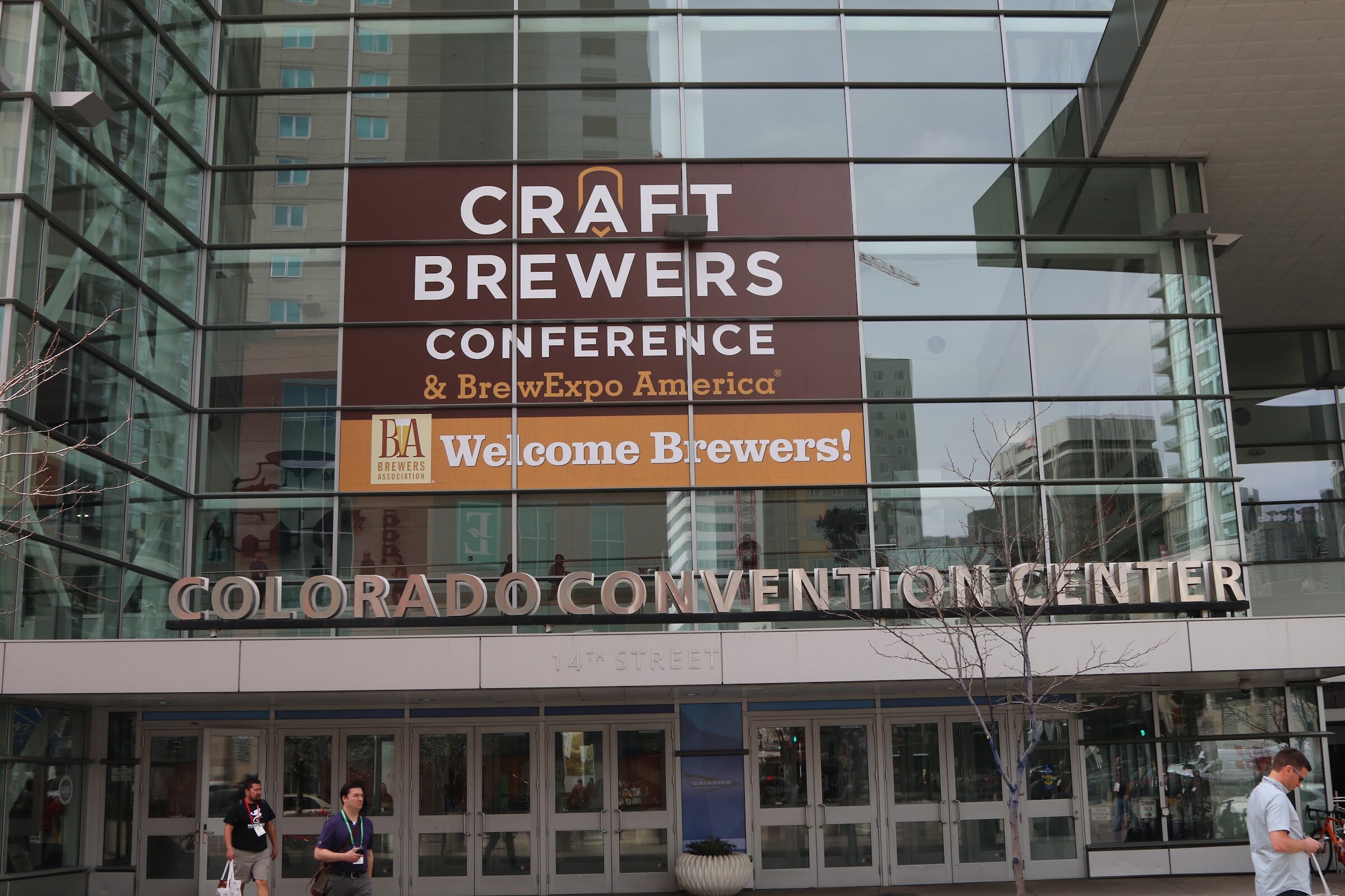Glass entrance of the Colorado Convention Center with a banner reading Craft Brewers Conference and BrewExpo America, Welcome Brewers.