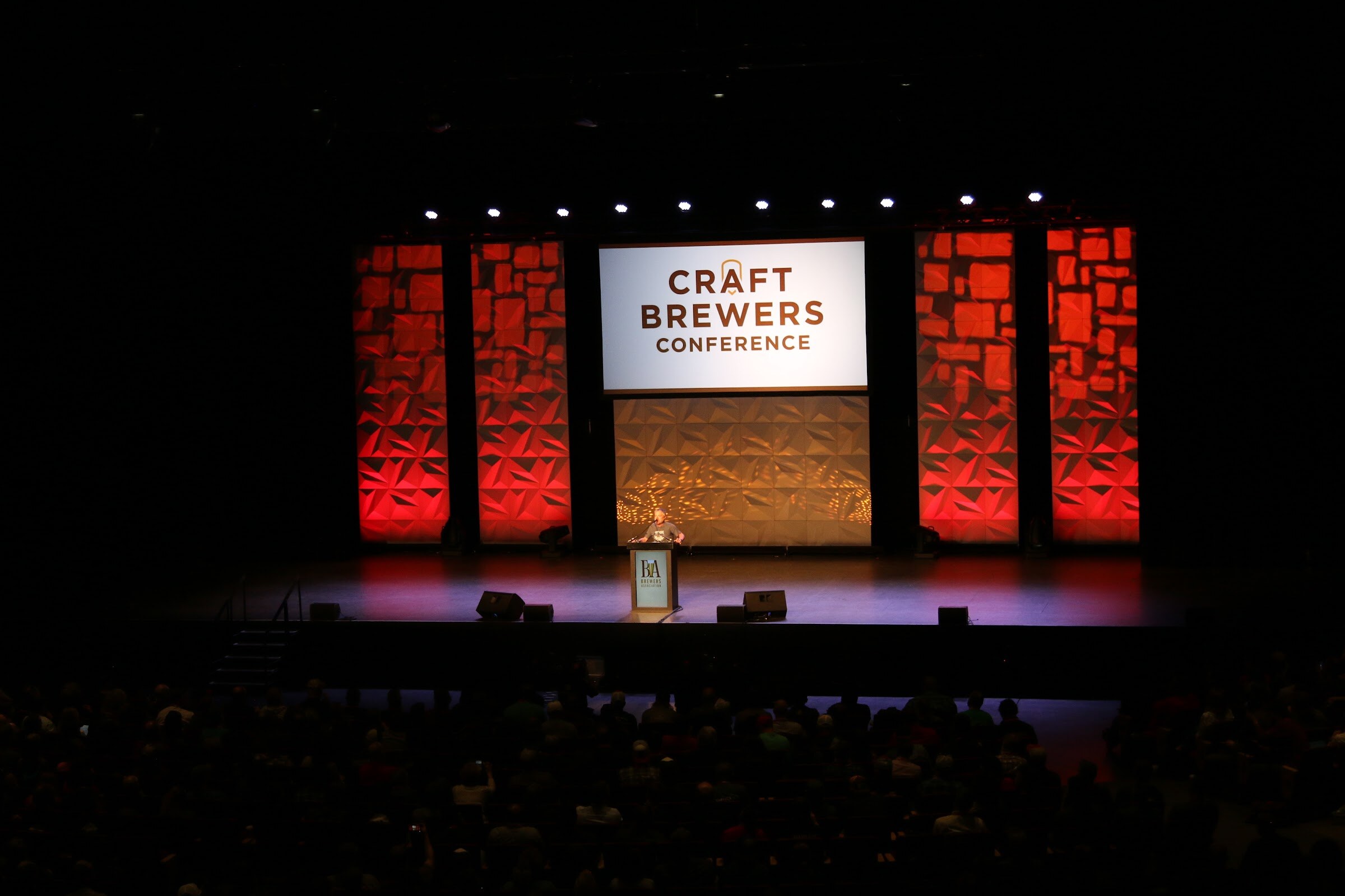 Wide view of a dark auditorium with a stage backdrop reading Craft Brewers Conference and a speaker at a podium.