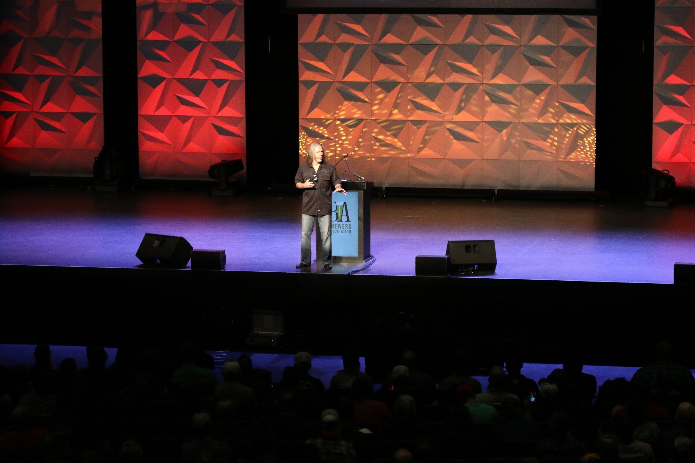 A man standing at a podium on a stage lit in blue and purple, with faceted orange backdrop panels behind.