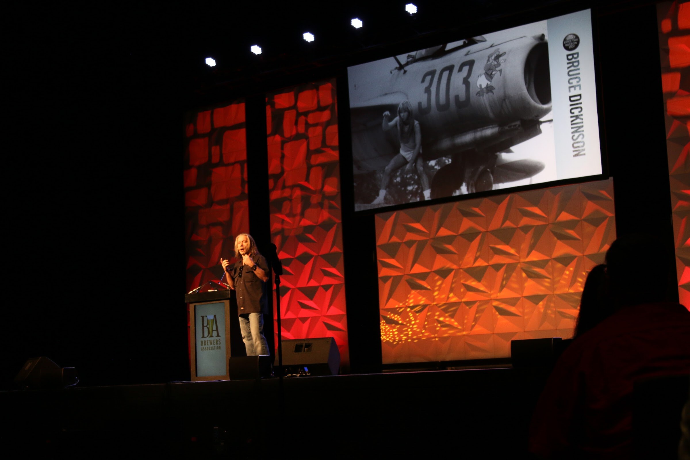 A speaker at a podium beside a large screen showing a black and white photo of an aircraft and the name Bruce Dickinson.